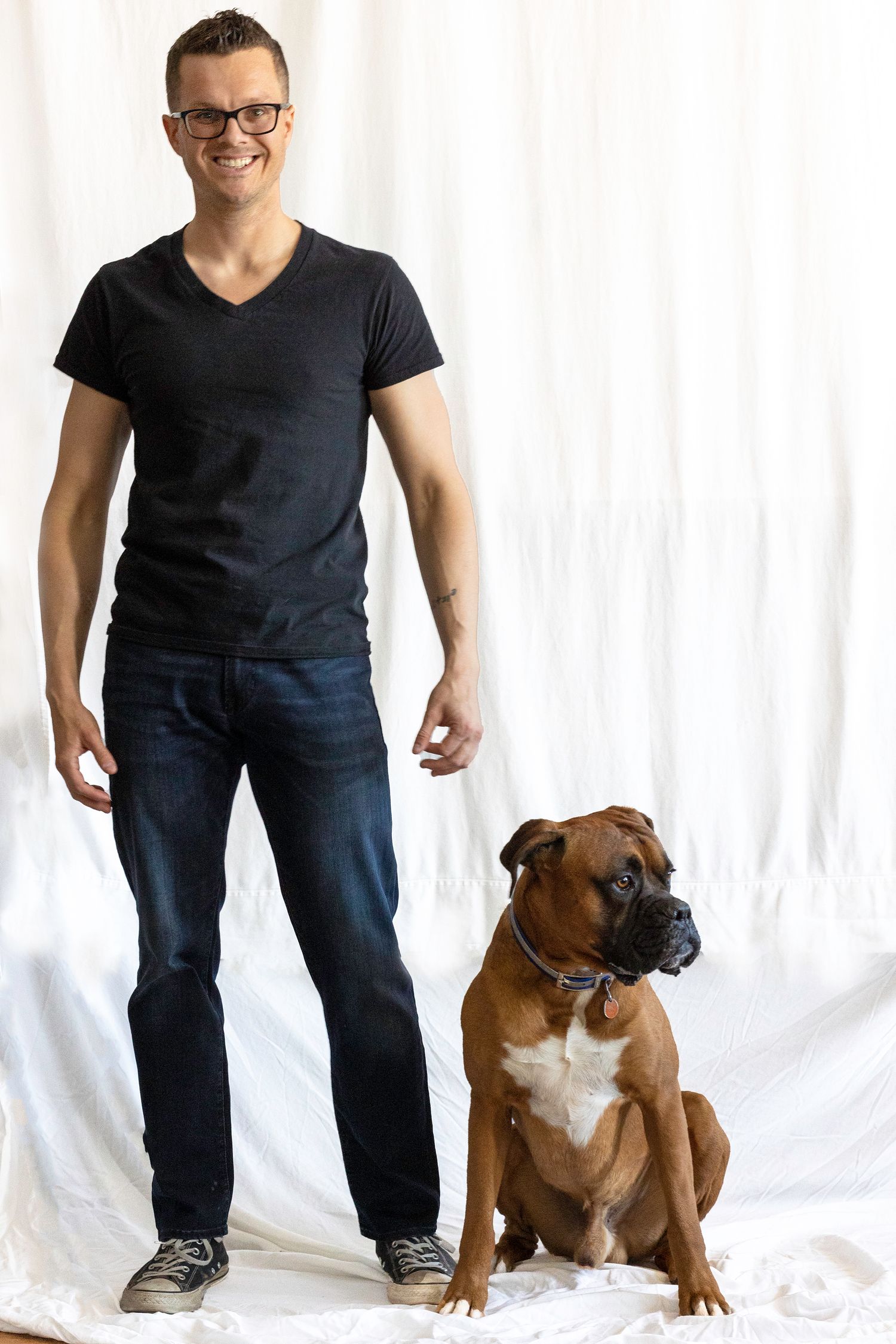 Smiling man with glasses standing next to fawn boxer dog against white backdrop.