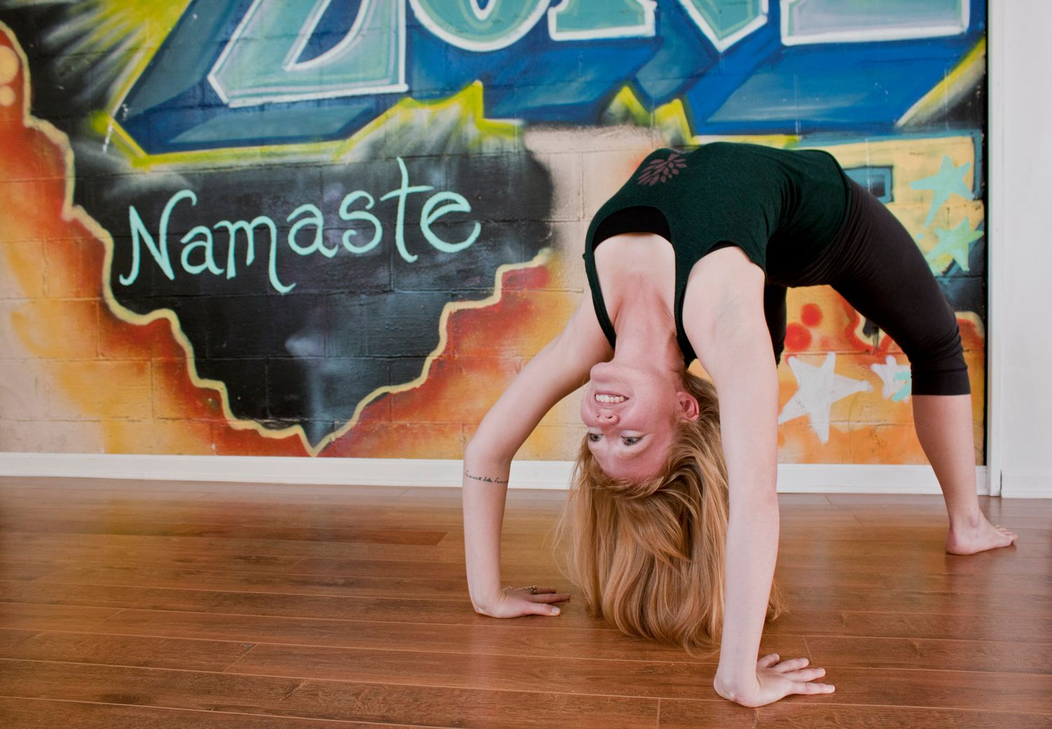 Woman performing a backbend yoga pose in front of a colorful Namaste graffiti mural on hardwood floors.
