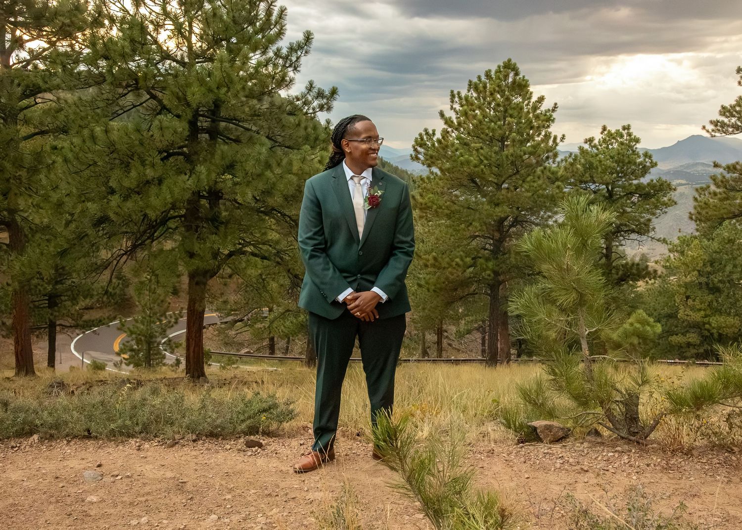 Man in green suit standing among pine trees with mountain backdrop in natural outdoor setting.