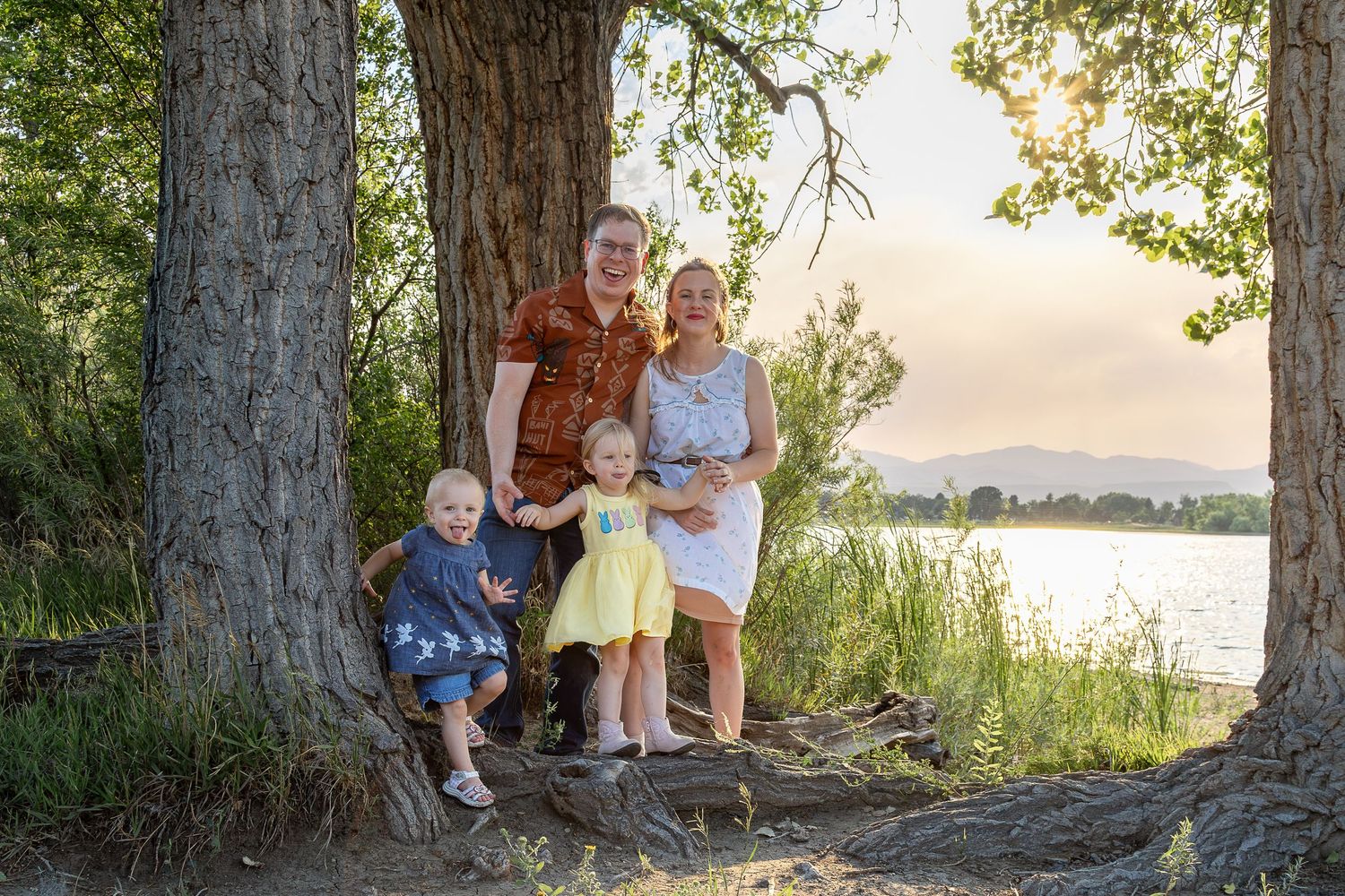 Family of four posing by large trees near a lake at sunset, two young children in colorful outfits smiling outdoors.