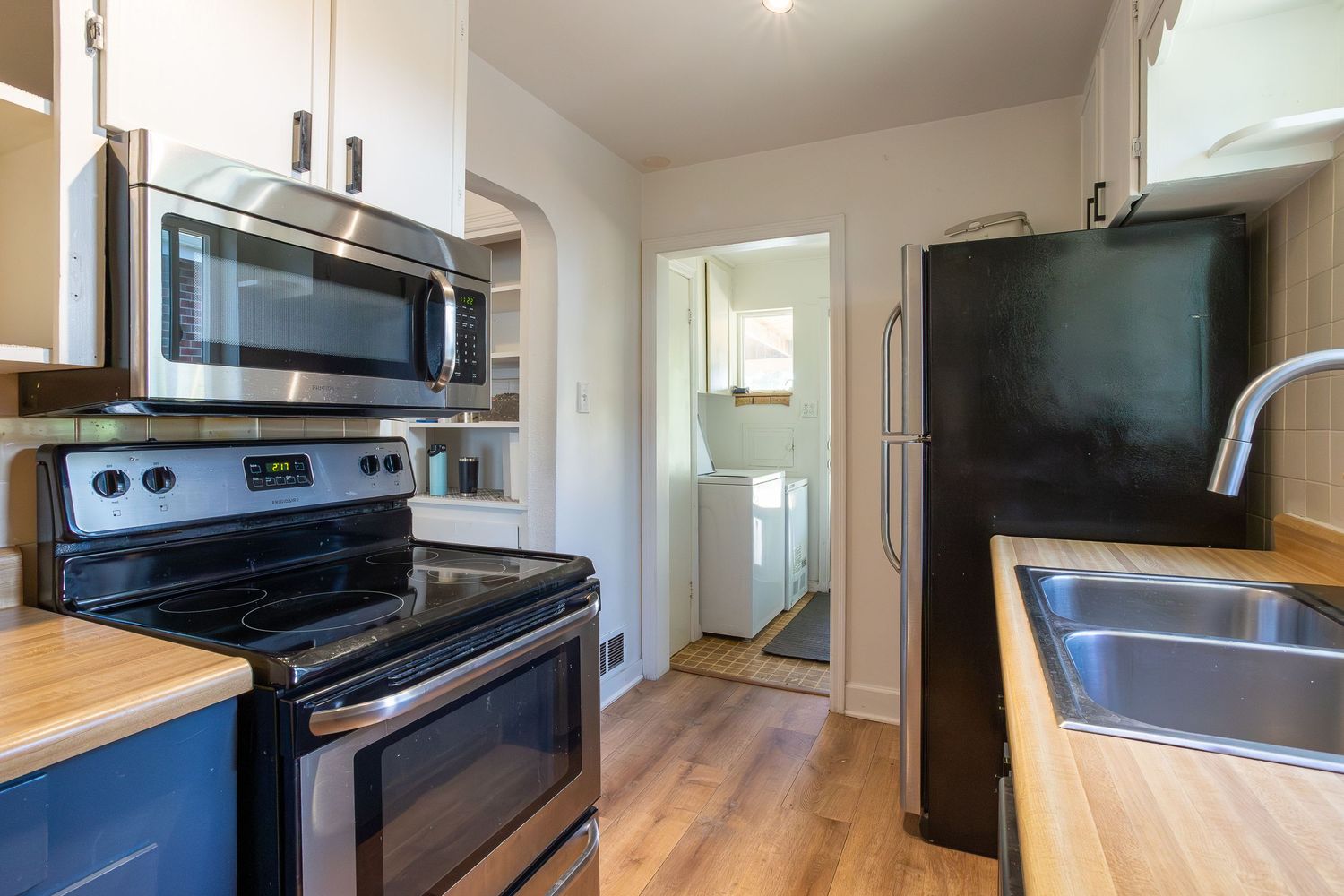 Modern kitchen with stainless steel appliances, butcher block countertops, hardwood floors, and a washer and dryer visible through doorway.