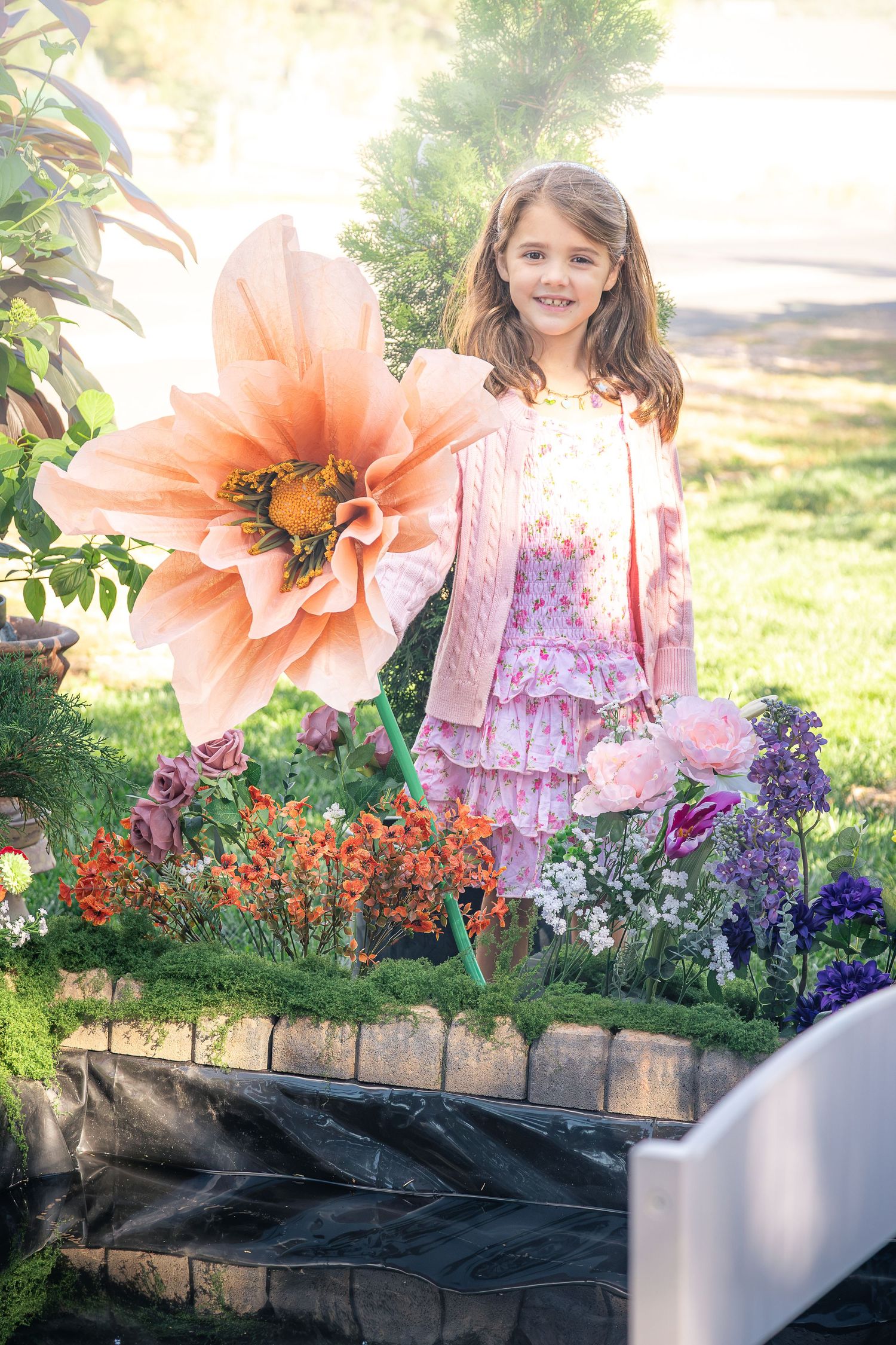 Smiling girl in a pink cardigan and floral dress stands beside a giant peach flower and colorful blooms in a sunny garden.