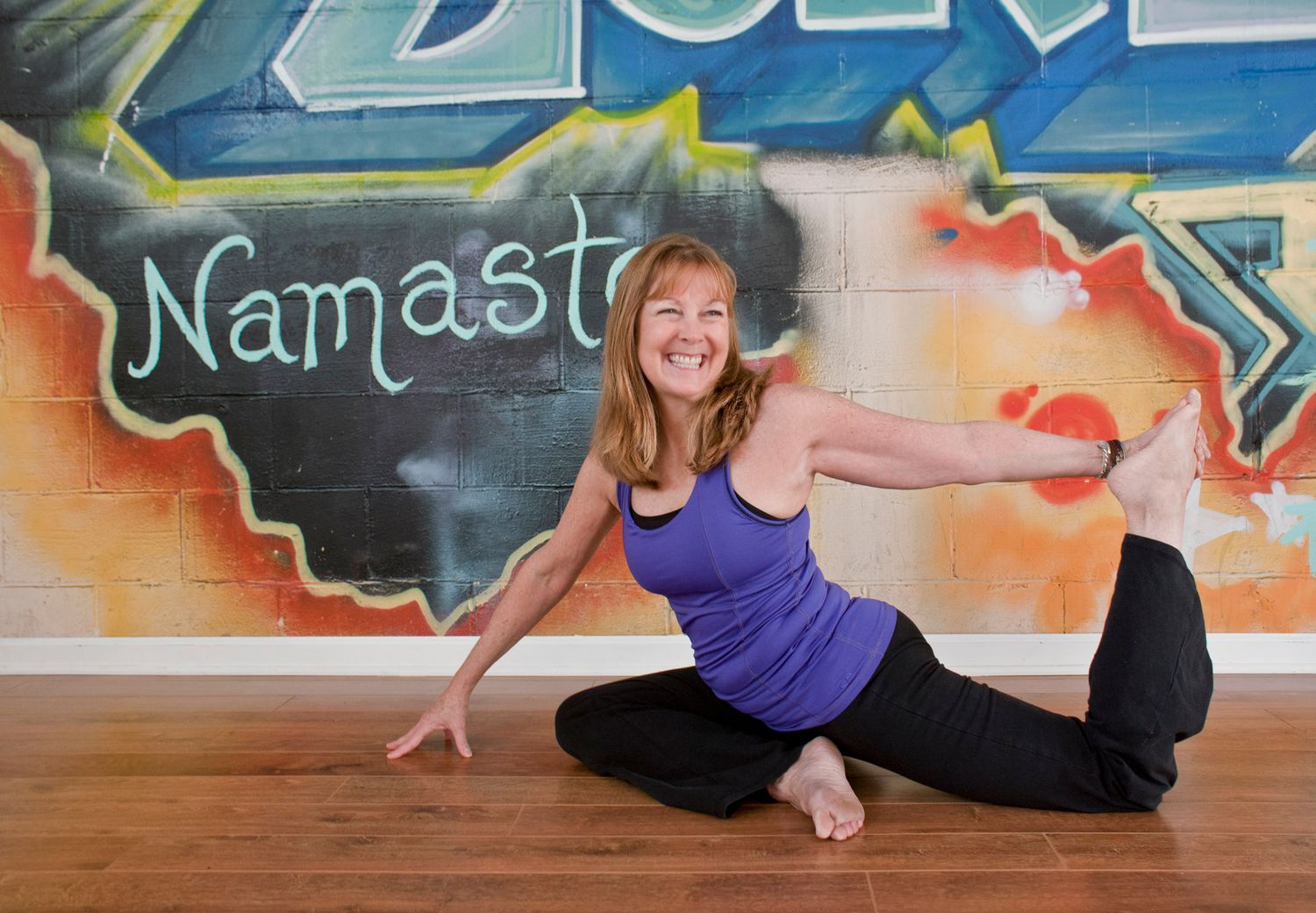 Smiling woman in purple tank performing seated yoga twist pose in front of Namaste graffiti wall.