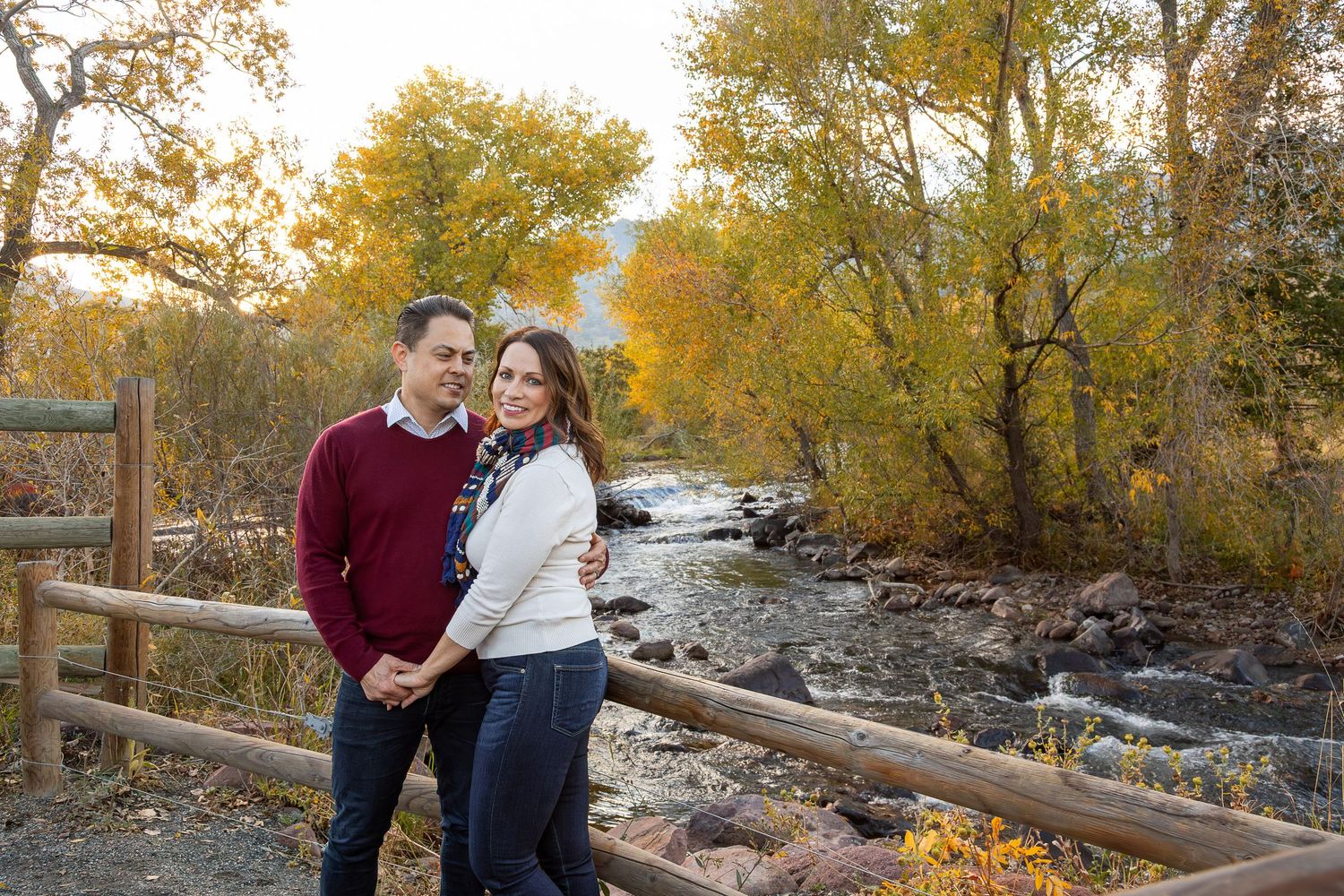 Couple embracing by a wooden fence near a rocky stream surrounded by colorful autumn foliage trees.