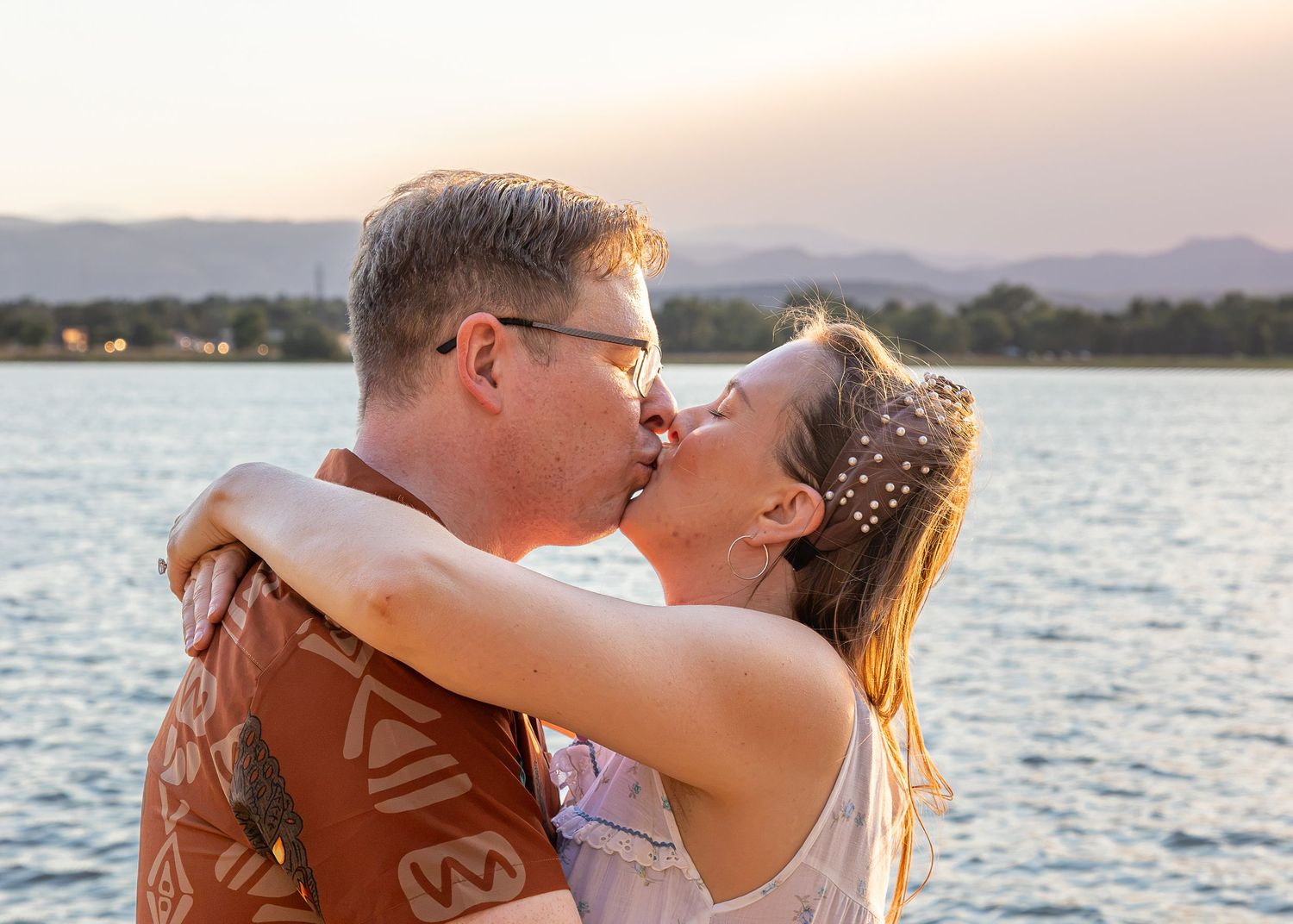 Couple sharing a romantic kiss by a lake with mountains at sunset, bride wearing pearl hair accessories.
