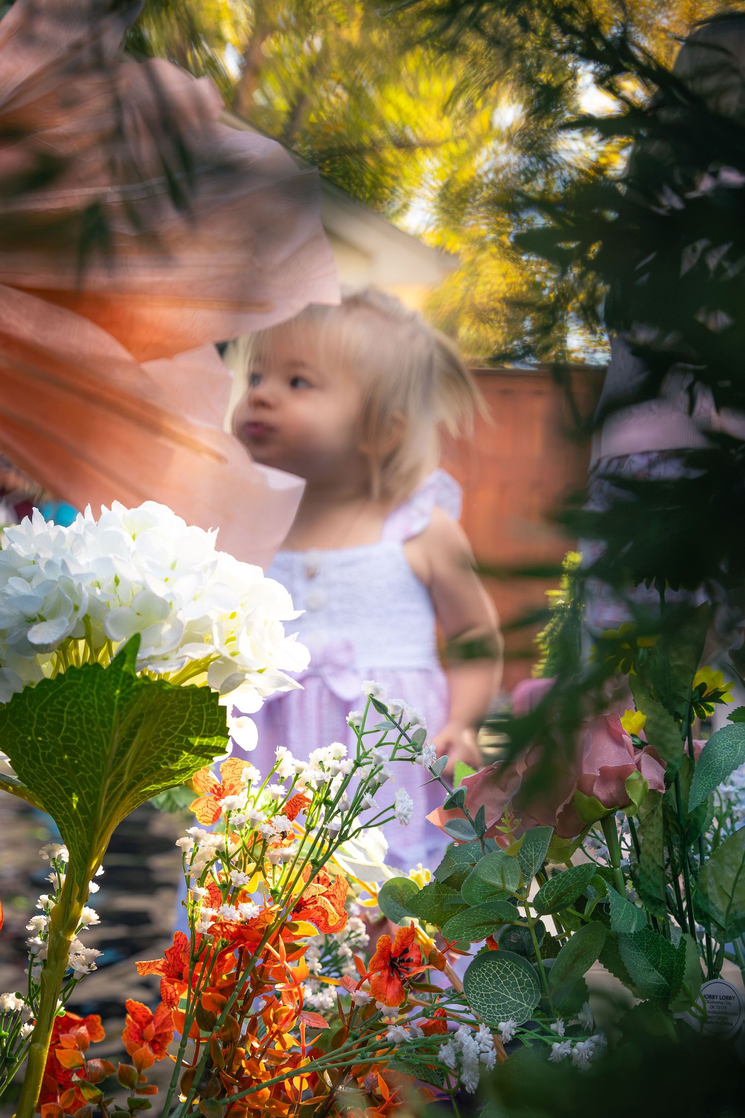 Blonde toddler in a lavender dress stands in a sunlit garden, framed by blurred white hydrangeas, orange flowers and green leaves.