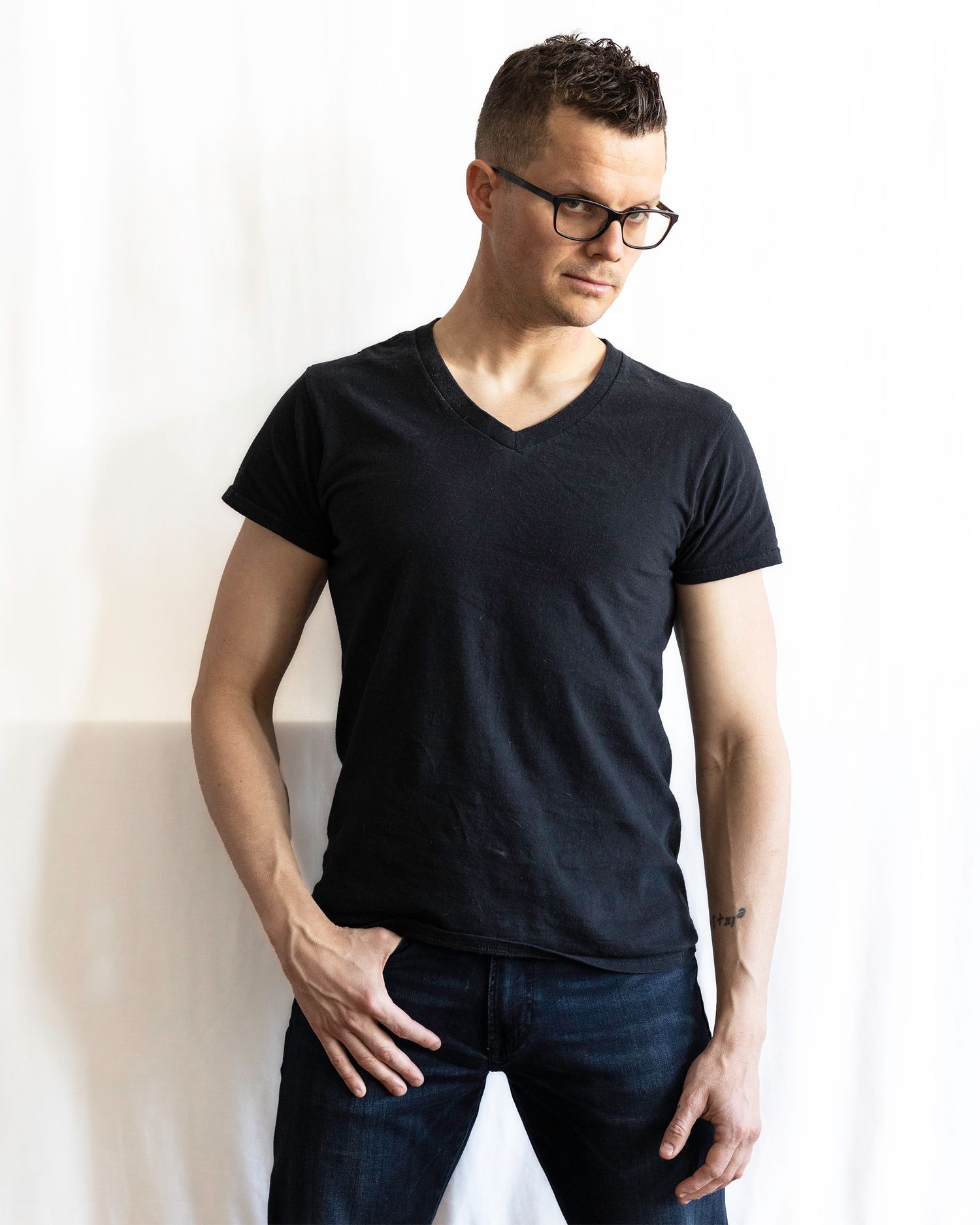 Slim man in black v-neck t-shirt and dark jeans leaning against a white curtain background.