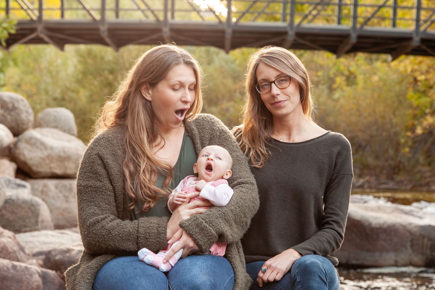 Two women sitting outdoors with a yawning baby, bridge and rocks in background.