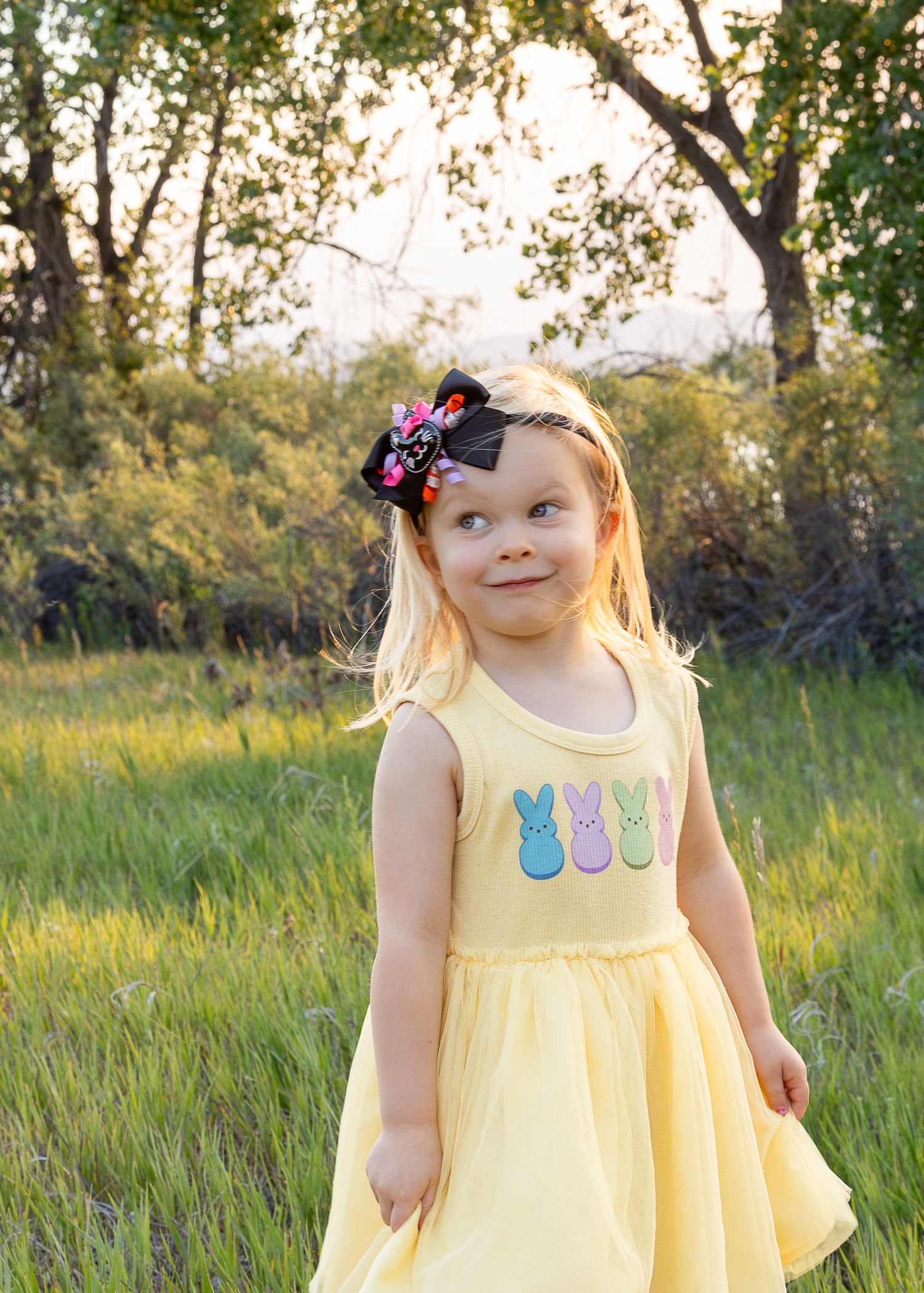 Blonde toddler girl in yellow Easter dress with Peeps design and black bow hair clip standing in green field.