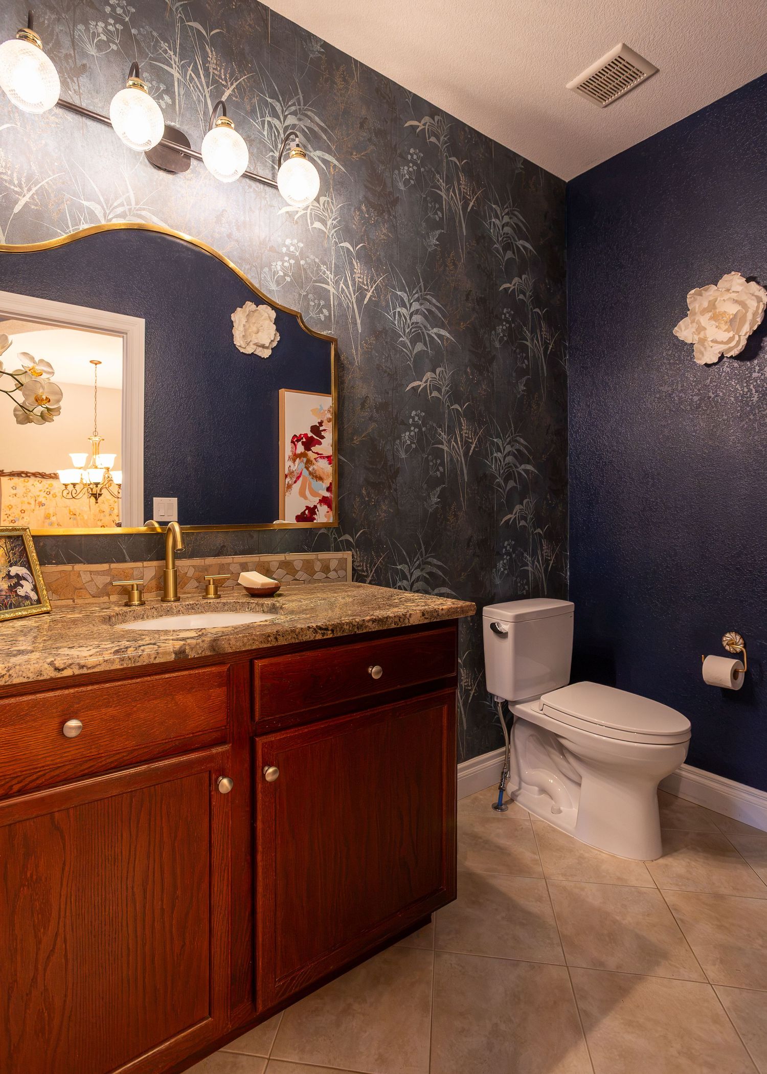 Elegant bathroom with navy floral wallpaper, cherry wood vanity, granite countertop, gold mirror, and white toilet.