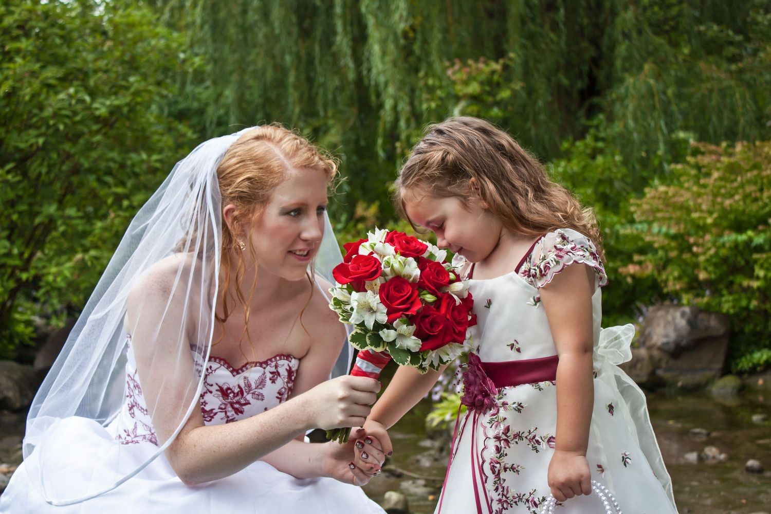 Bride in white gown with red floral details shares bouquet of red roses with young flower girl outdoors.