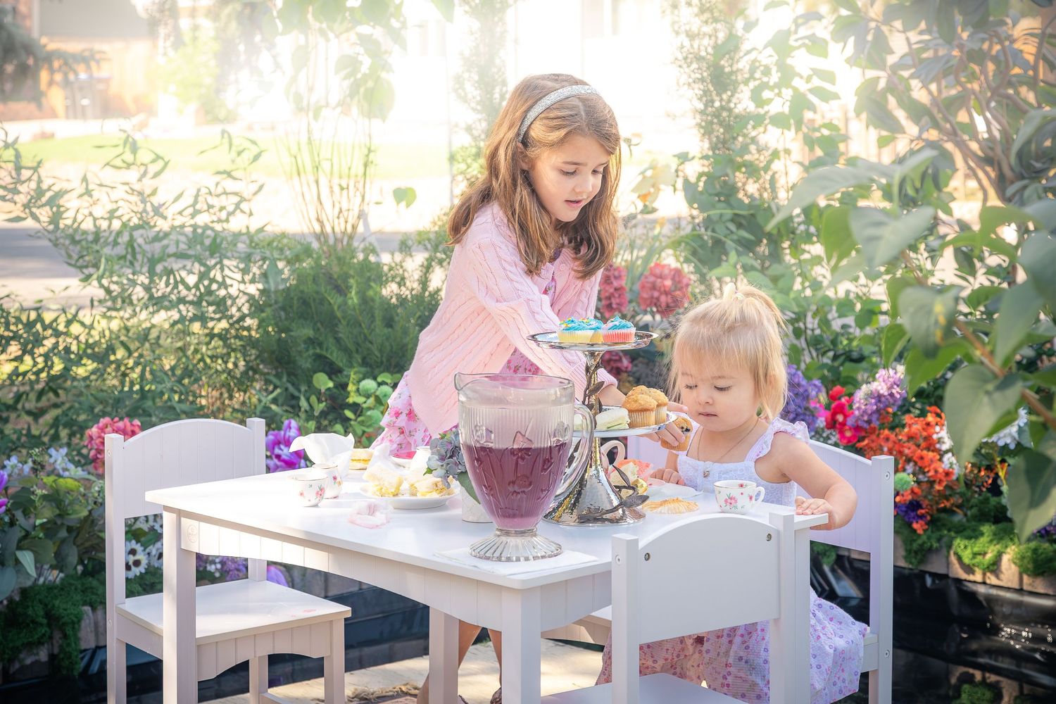 Two young girls enjoy a tea party at a white table with a glass pitcher of berry juice and snacks, surrounded by blooming flowers.