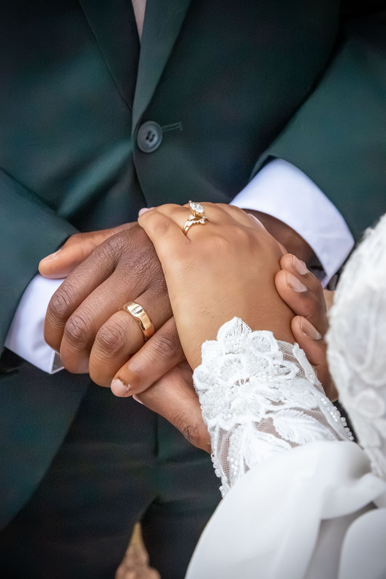 Interracial newlywed couple holding hands, wearing gold wedding rings, groom in green suit, bride in white lace dress.