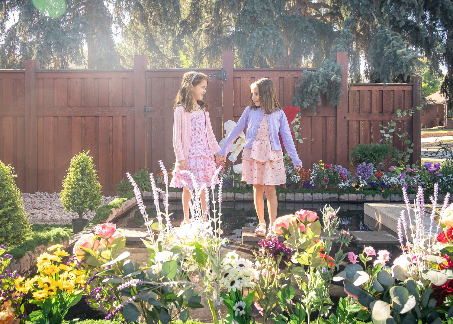 Two young girls in pastel dresses hold hands while standing in a sunlit backyard garden with colorful blooming flowers and a wooden fence.