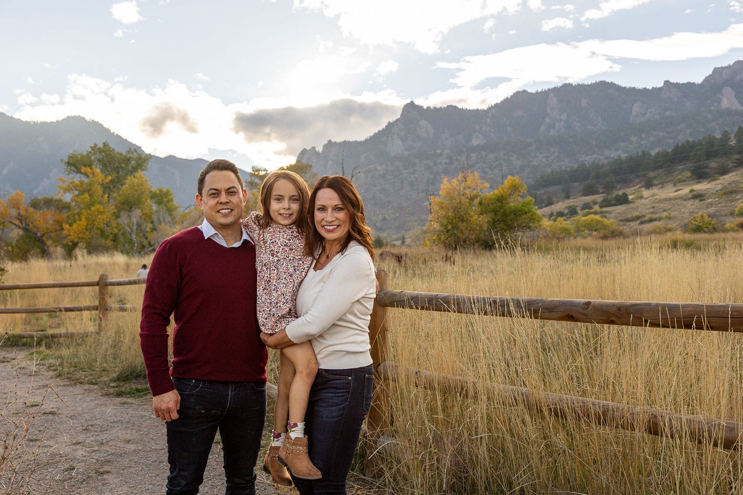 Family of three posing outdoors near wooden fence with Rocky Mountain landscape and golden fields in background.