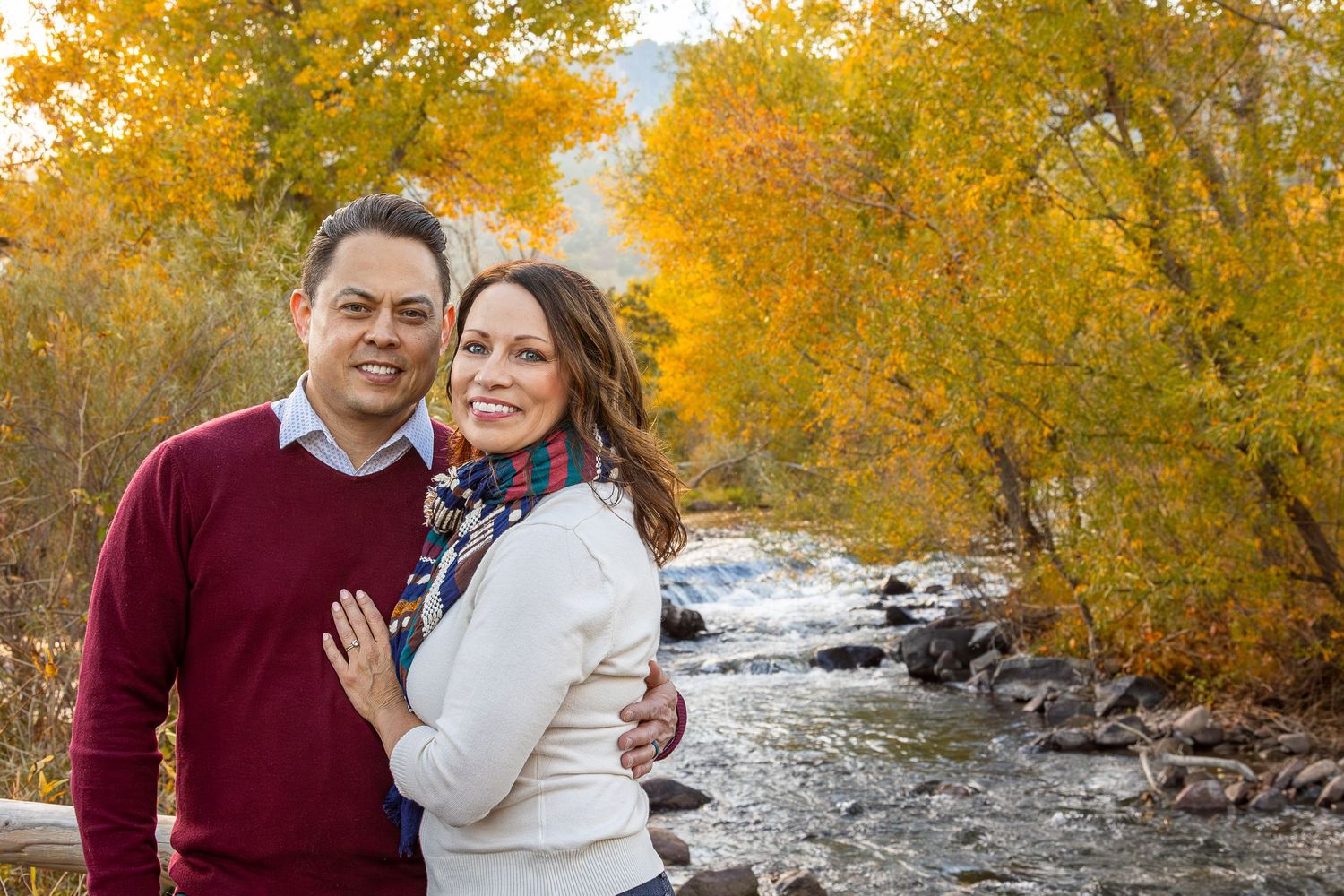 Couple smiling together outdoors by a rocky stream surrounded by vibrant yellow autumn foliage trees.