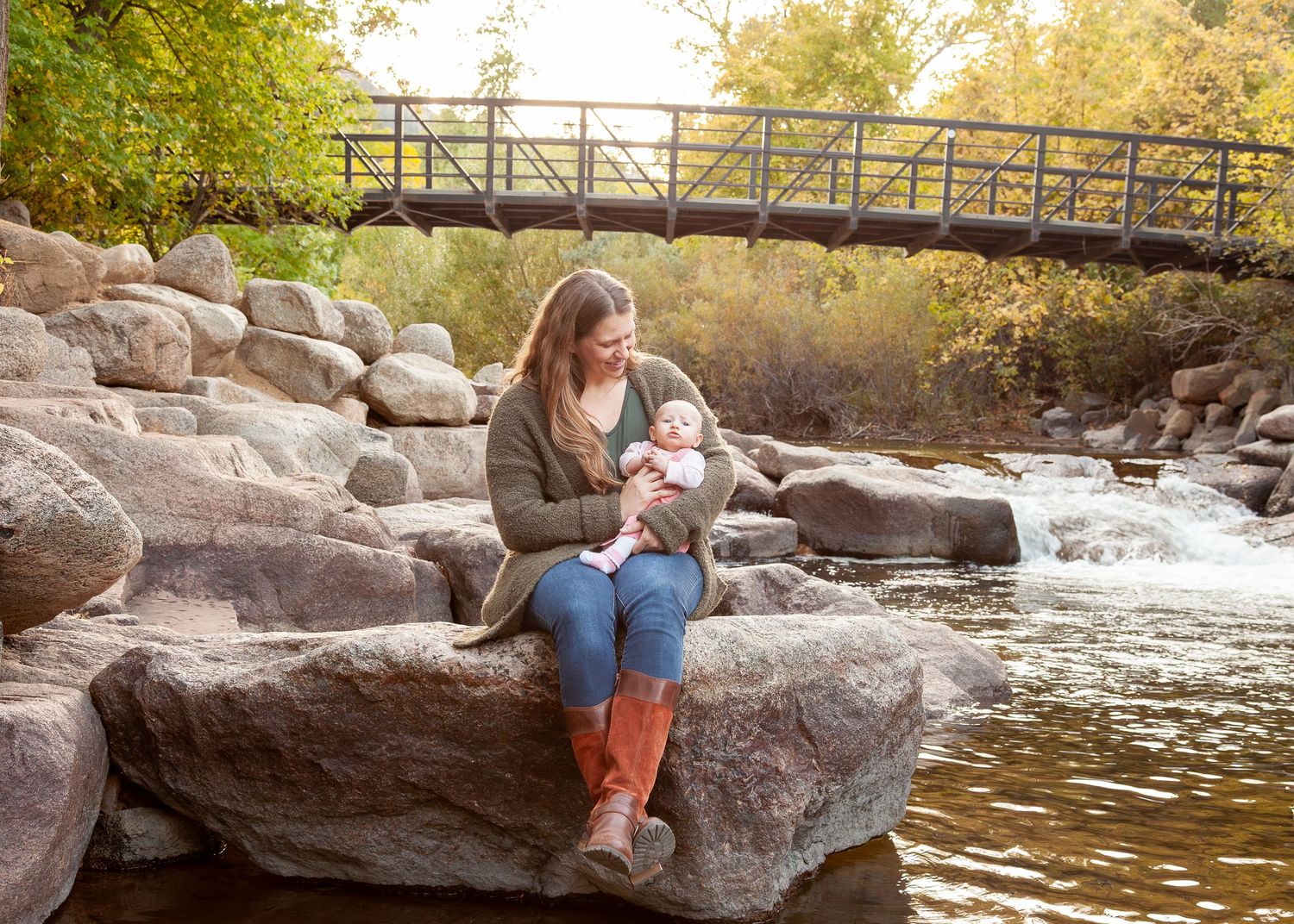 Mother sitting on rocks by a creek holding her baby, with a bridge and autumn trees in the background.