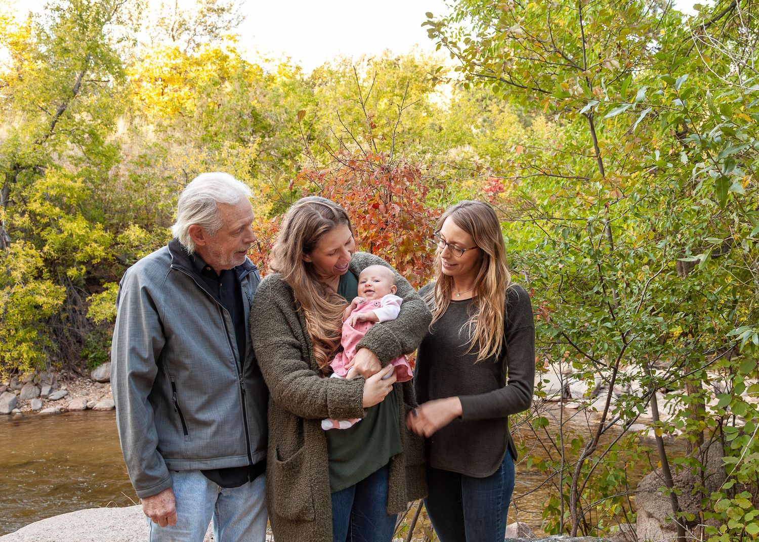 Multi-generational family admiring newborn baby outdoors near a river surrounded by colorful autumn foliage.