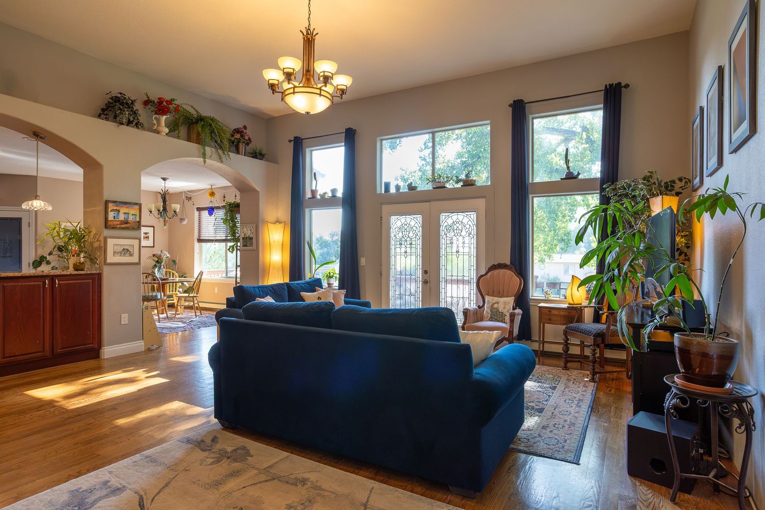 Bright living room with blue sofas, hardwood floors, tall windows, chandelier, and lush indoor plants in an elegant home.