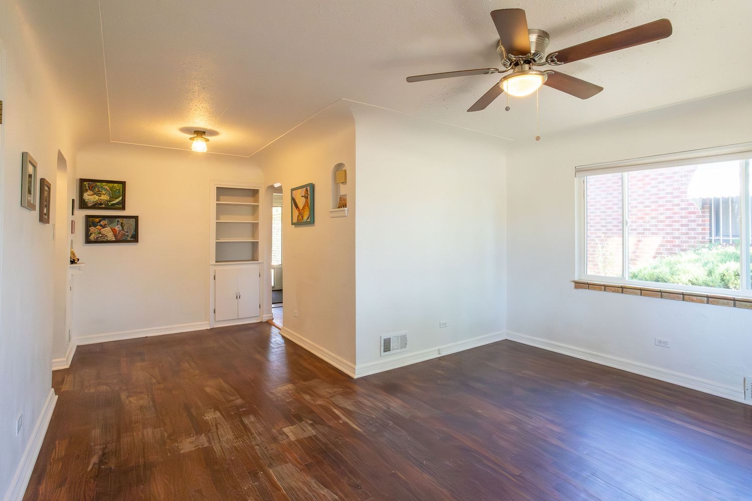 Spacious living room with dark hardwood floors, ceiling fan, built-in shelving, and natural light from a large window.
