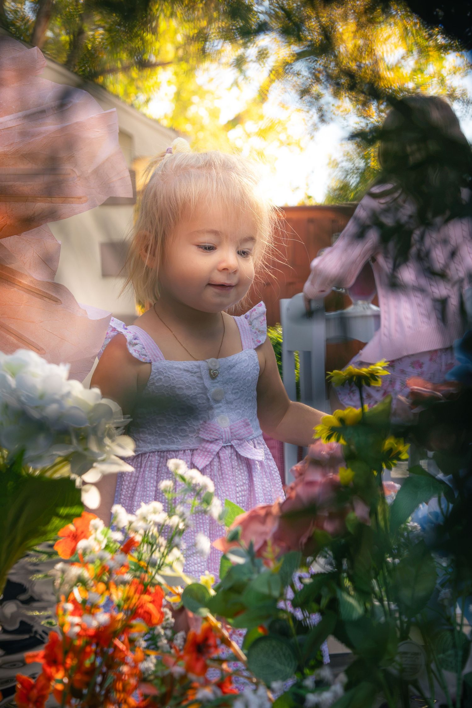 Smiling toddler girl in a lavender dress stands among colorful orange, pink and white flowers in a sunlit garden.