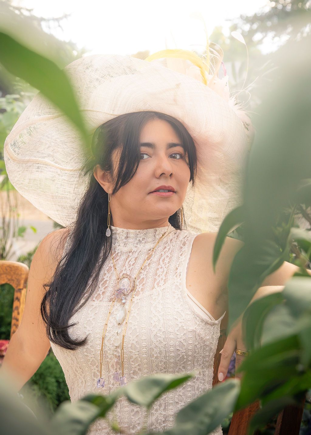 Woman with long dark hair wears a wide-brimmed cream hat and lace sleeveless dress with layered necklaces, framed by green garden leaves.