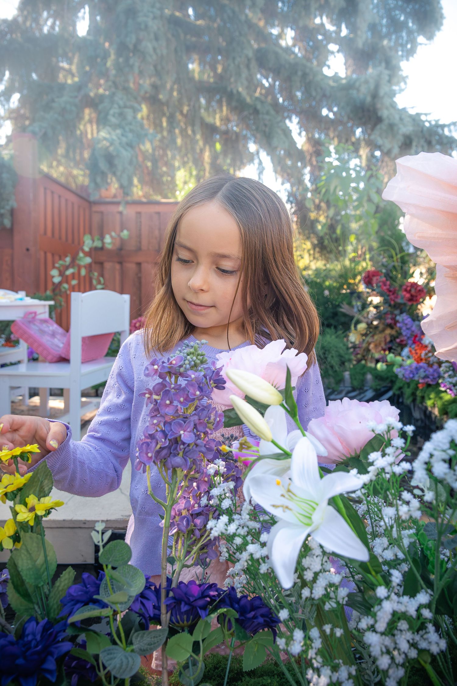 Young girl with long brown hair admires pink roses, white lilies, and purple delphiniums in a colorful garden with a wooden fence.