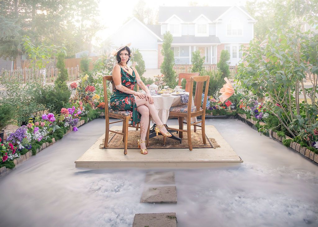 Woman in a floral dress sits at a small bistro table on a wooden patio surrounded by colorful flower beds, with a house in the background.