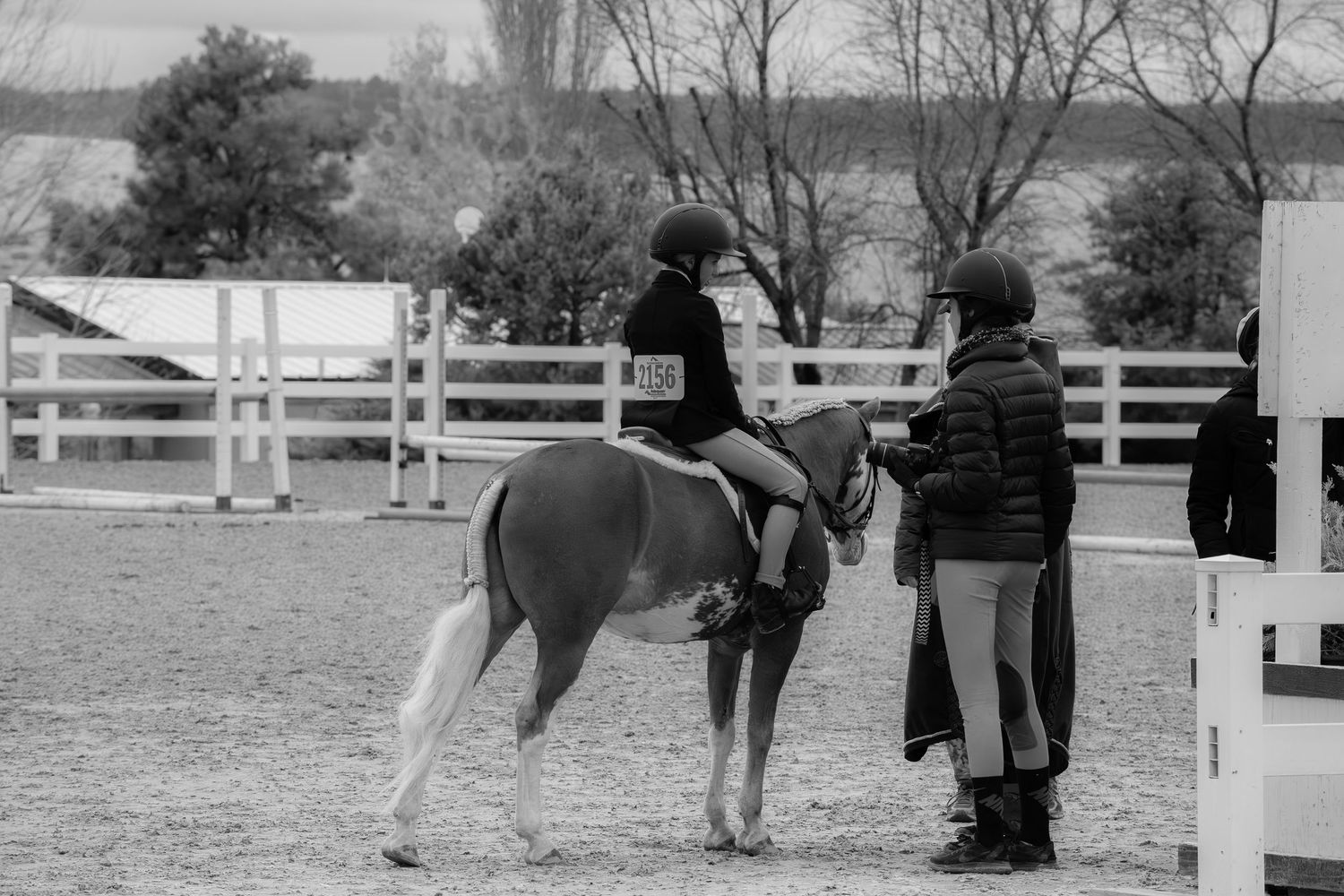 Young rider on a pony receives instruction from a trainer at an equestrian arena, black and white.