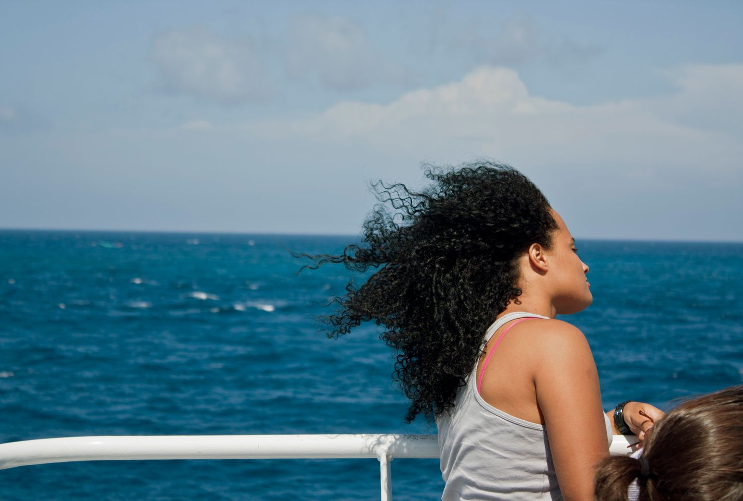 A woman with curly black hair enjoys ocean breeze on a boat deck under blue skies.