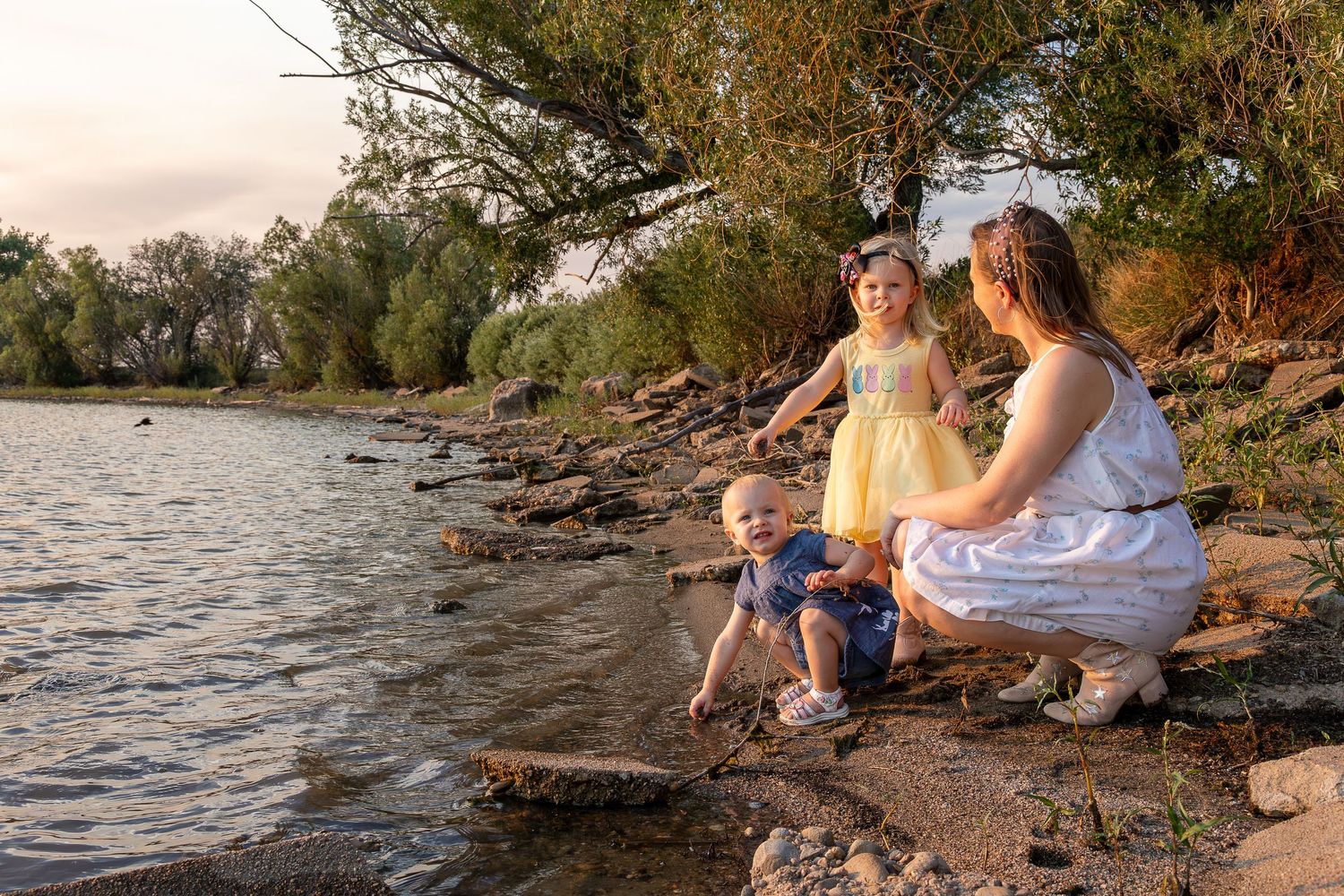 A mother and two young children playing by a rocky riverbank surrounded by lush green trees at sunset.