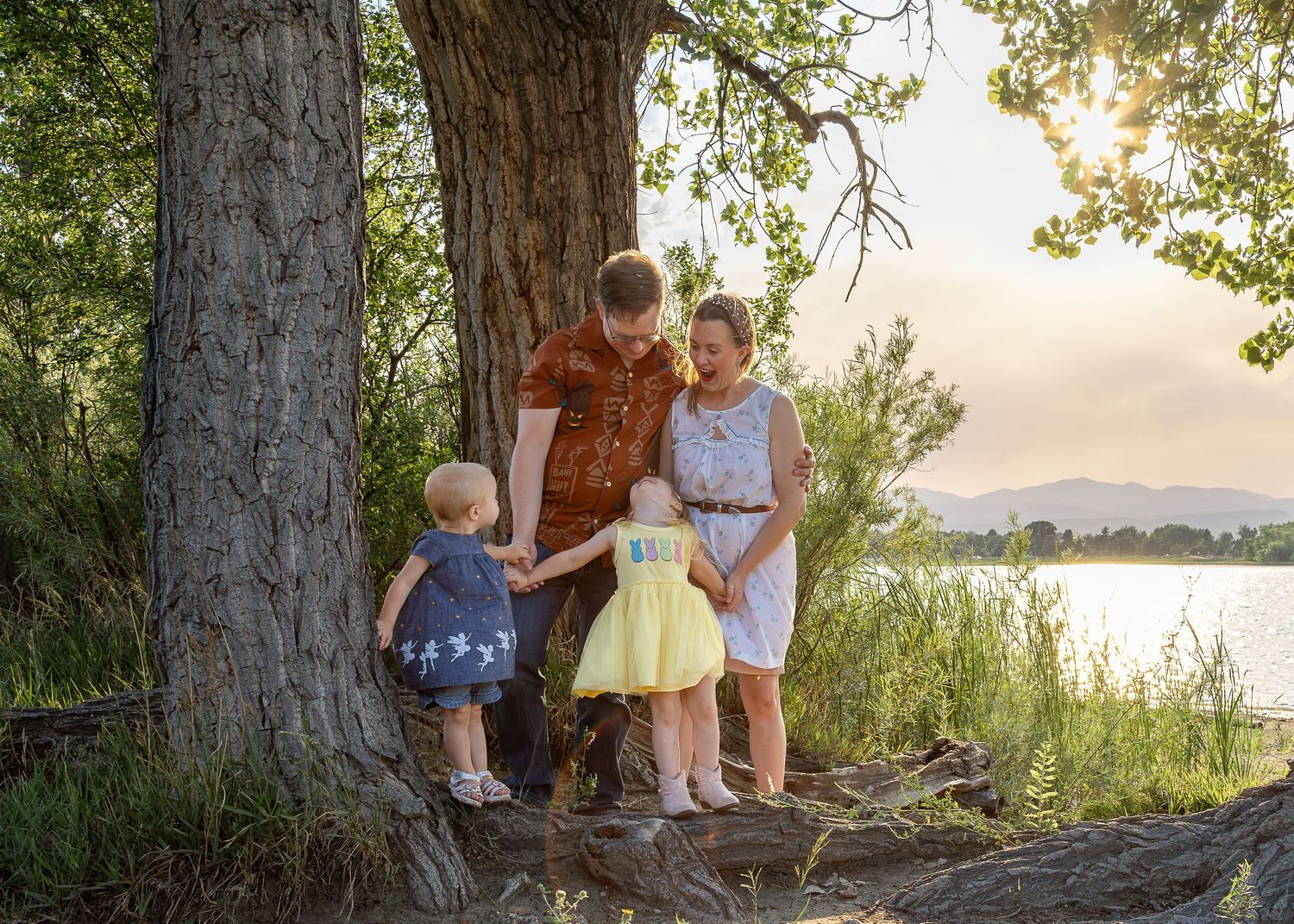 Family of four standing by a large tree near a lake at sunset, parents watching their two young children.