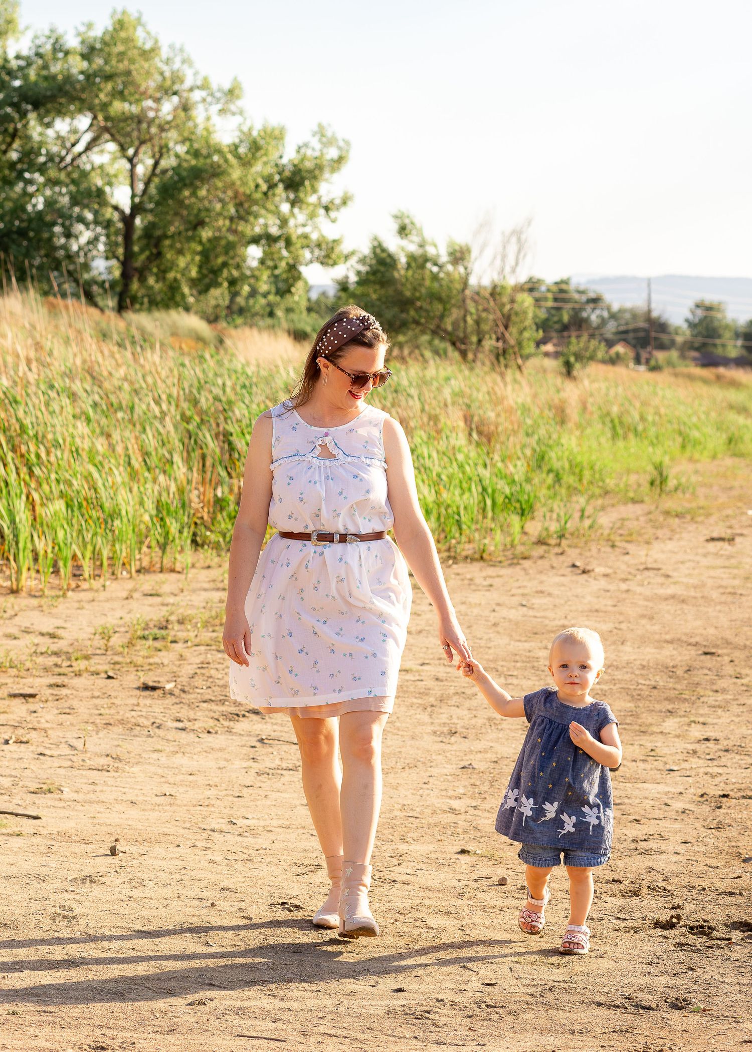 A mother in a white dress holds hands with her toddler in a blue dress while walking on a dirt path.