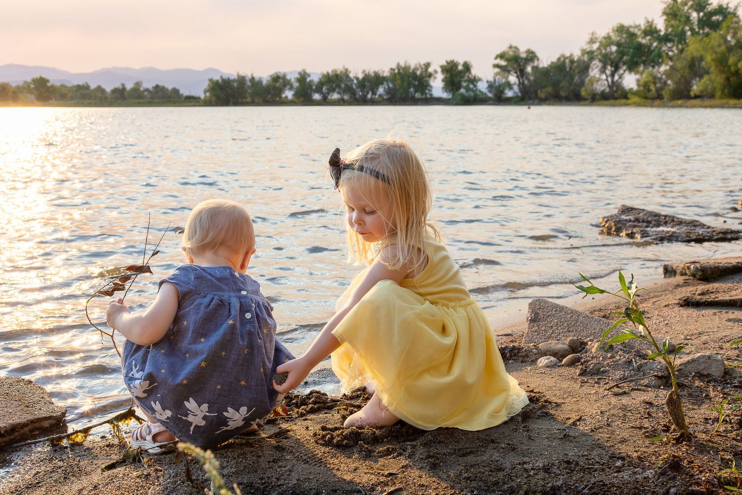 Two young children sitting on a sandy lakeshore at sunset, playing together near calm water with trees in background.