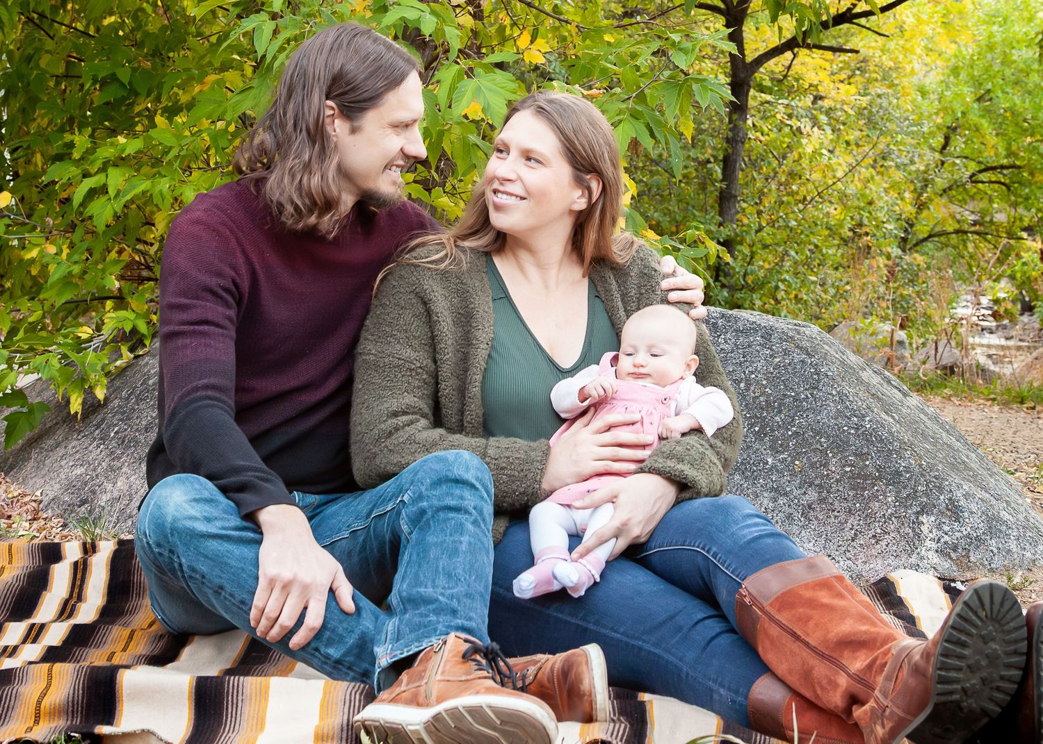 A couple sits on a blanket against rocks, smiling at each other while holding a newborn baby outdoors in autumn.
