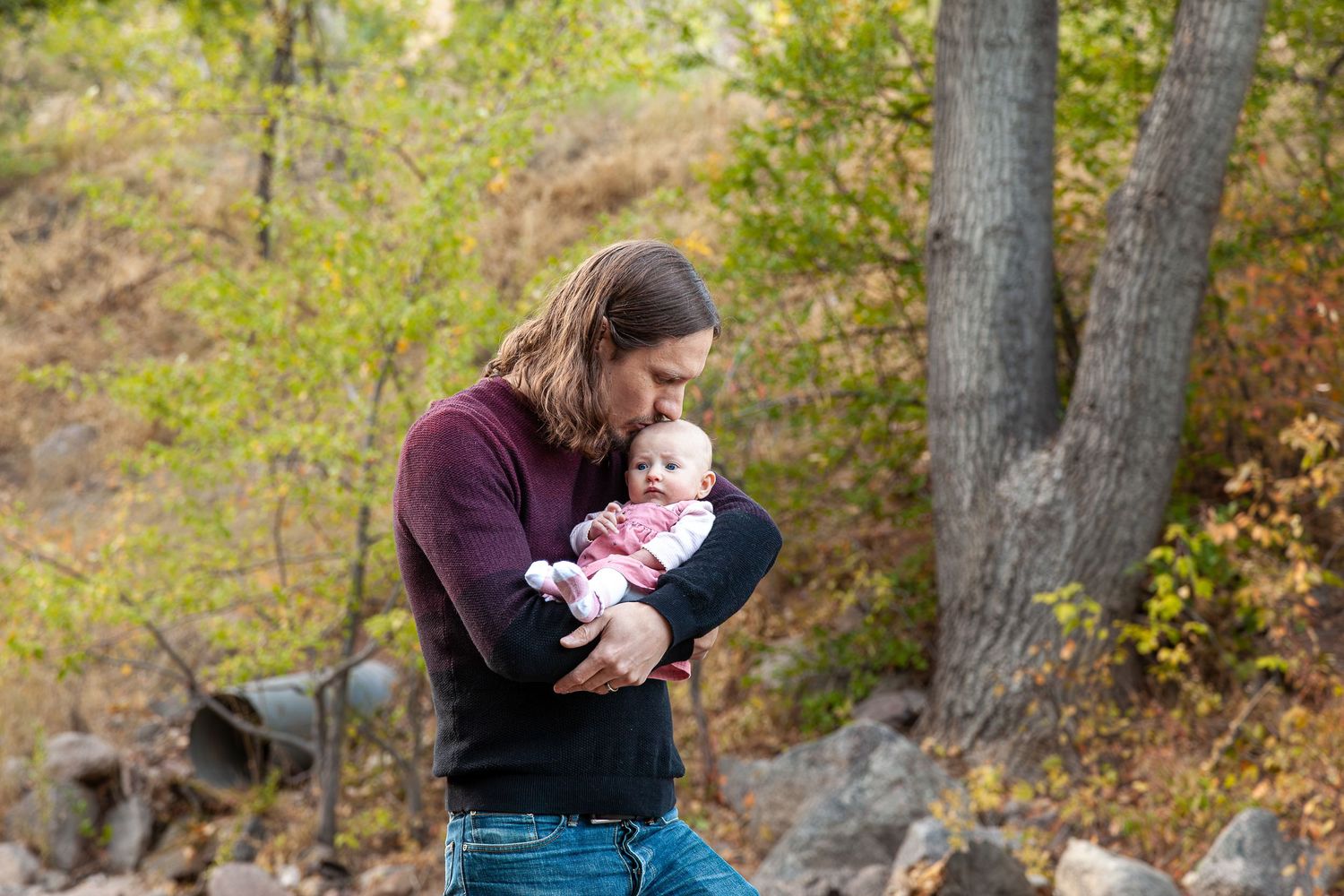 A man with long hair kisses a newborn baby outdoors in a forest setting with autumn foliage.
