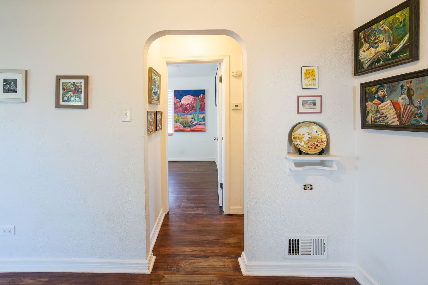 Hallway with arched doorway, hardwood floors, white walls adorned with colorful framed artwork, and a decorative plate on a shelf.
