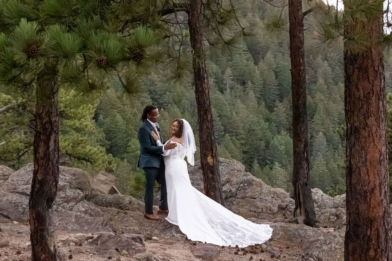 Bride and groom share a romantic moment among pine trees on rocky terrain with lush green forest backdrop.