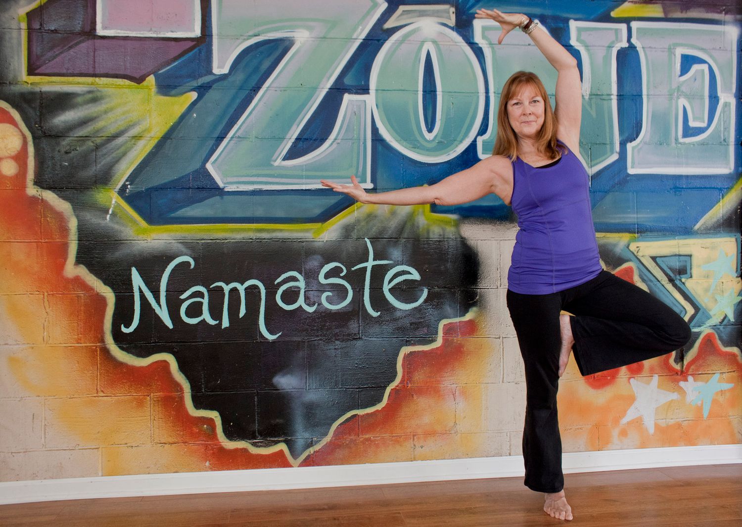 Woman in purple tank top performing standing balance yoga pose in front of Namaste graffiti wall.