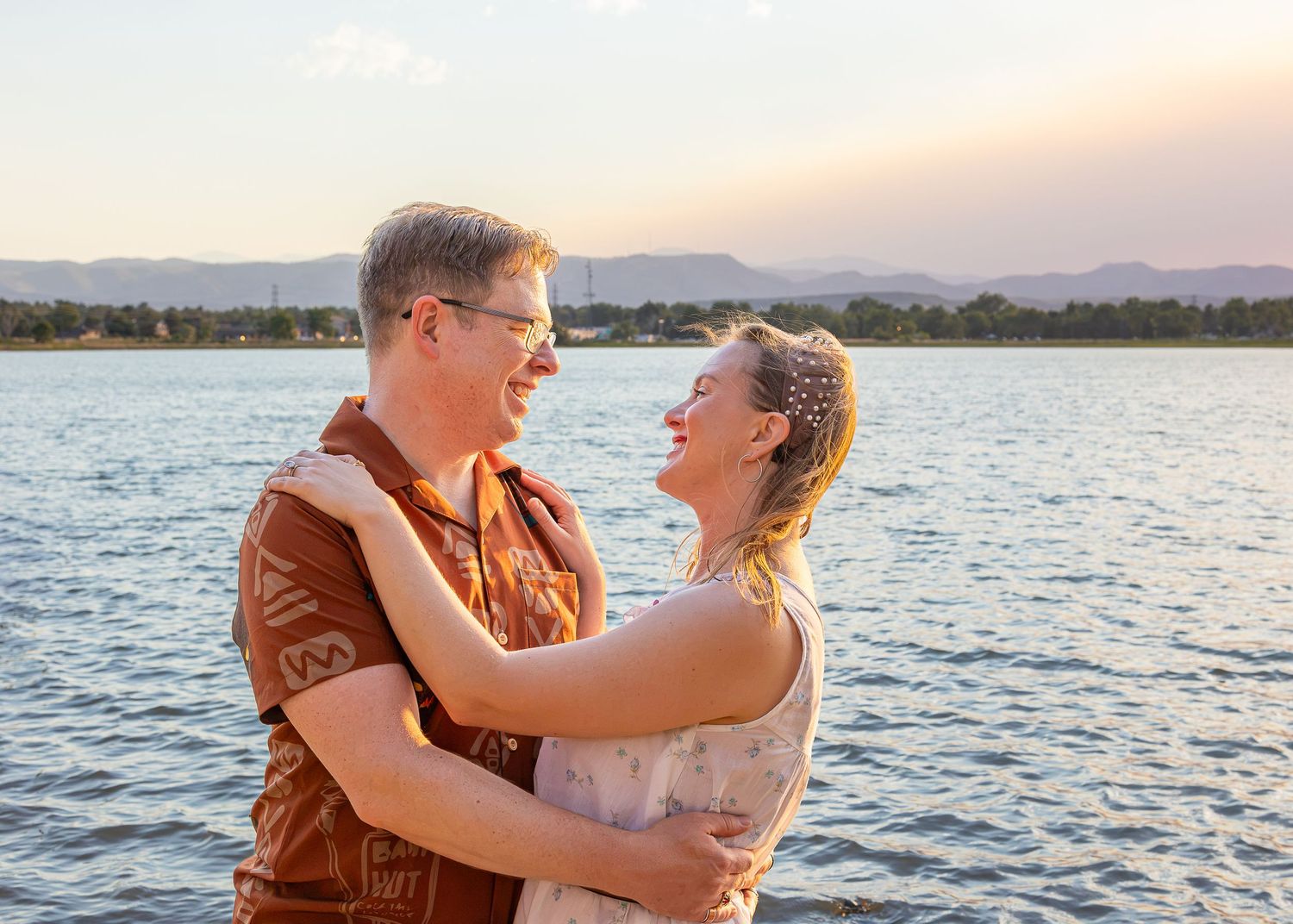 Couple embracing and smiling by a lake at sunset with mountains in the background.