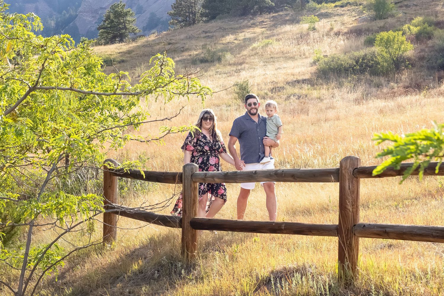 Family of three posing by a rustic wooden fence in a sunny golden meadow with green hills and trees in the background.
