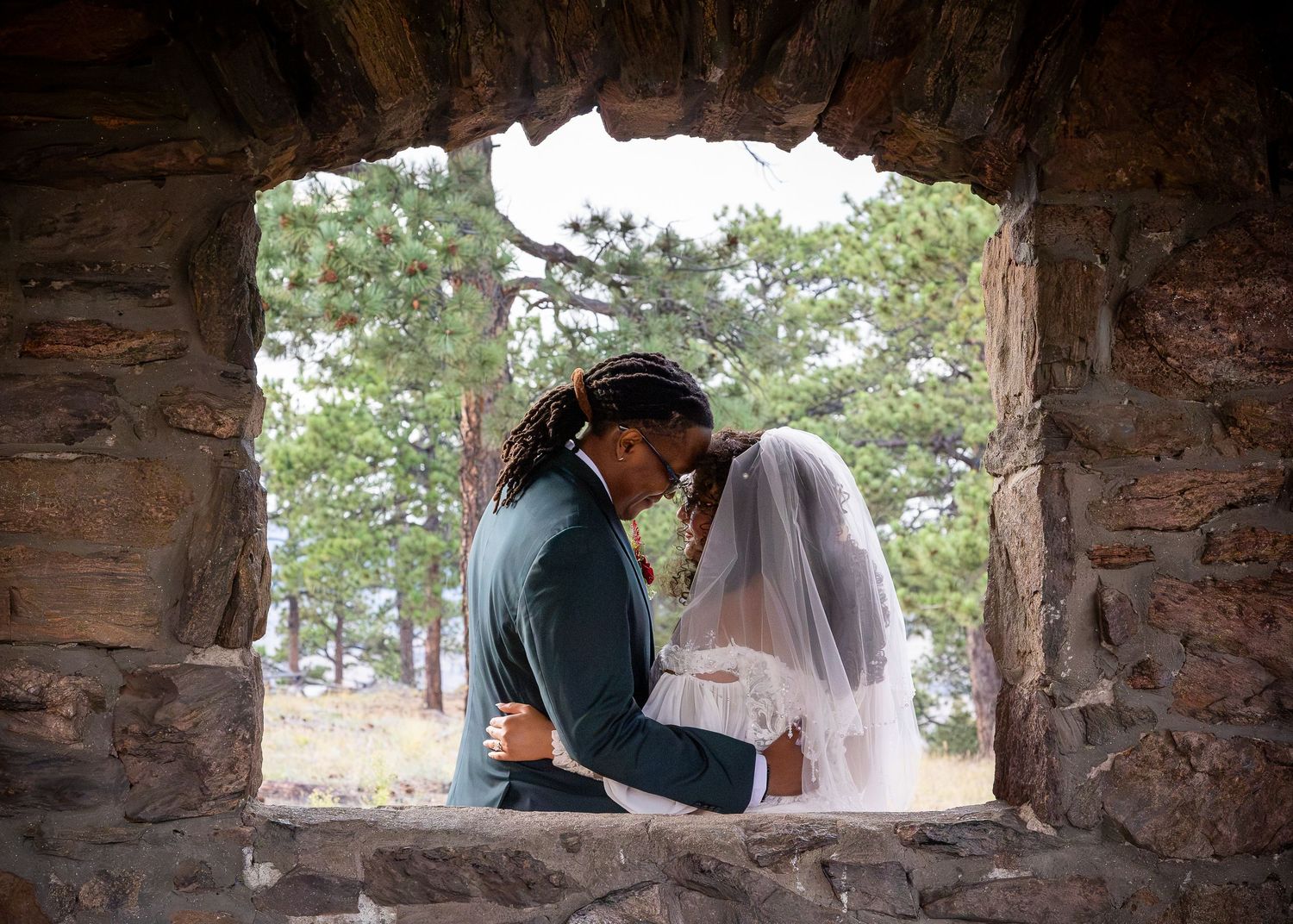 Newlyweds share an intimate moment framed by a rustic stone window arch outdoors.