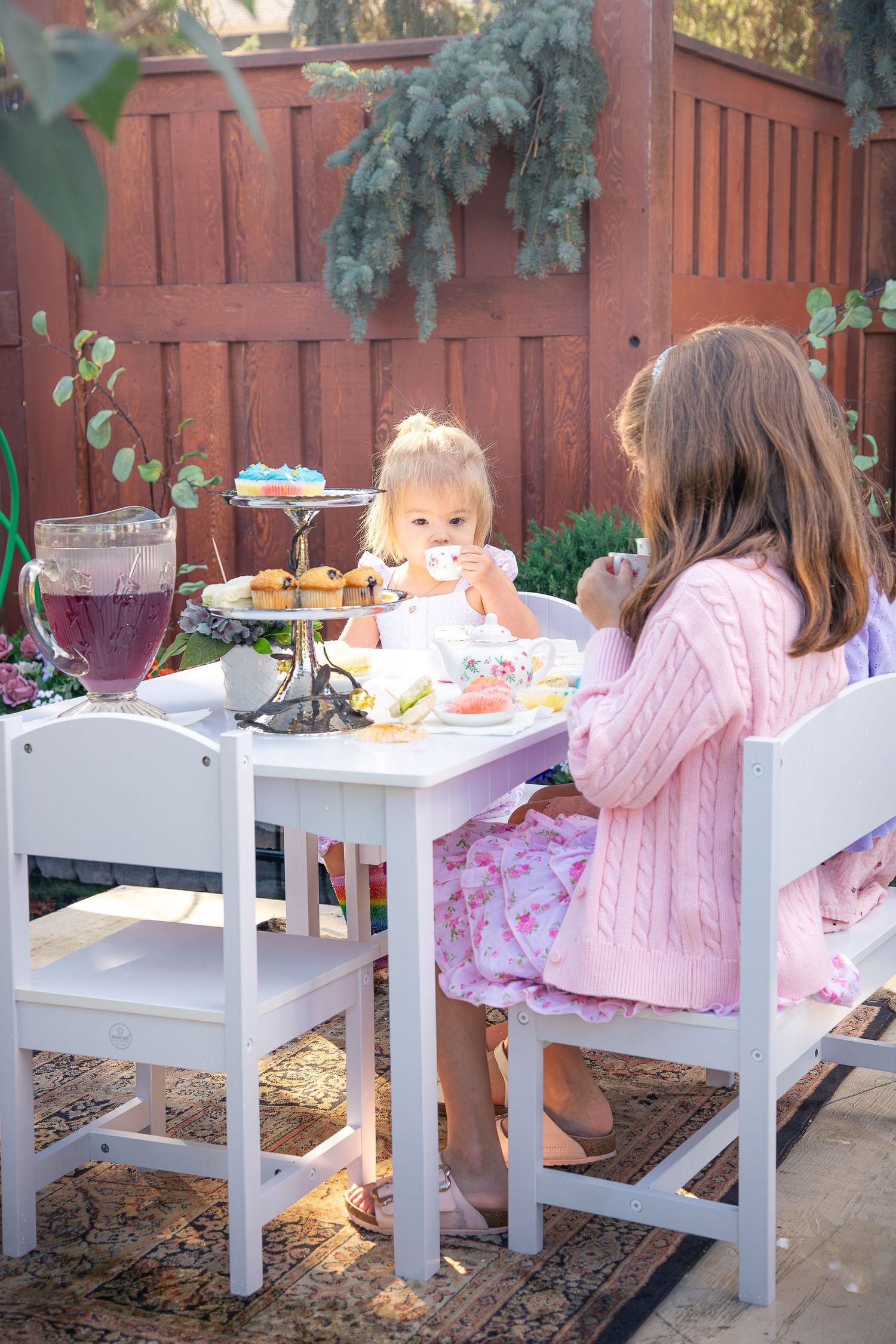 Two young girls enjoy a tea party at a white child-sized table outdoors with a tiered tray of treats and a pitcher of berry drink.