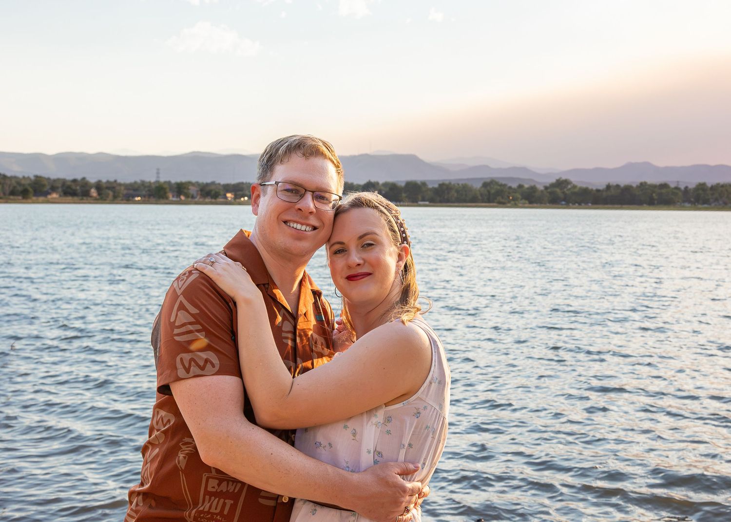 Couple embracing and smiling by a lake with mountains at sunset, romantic outdoor portrait photography.