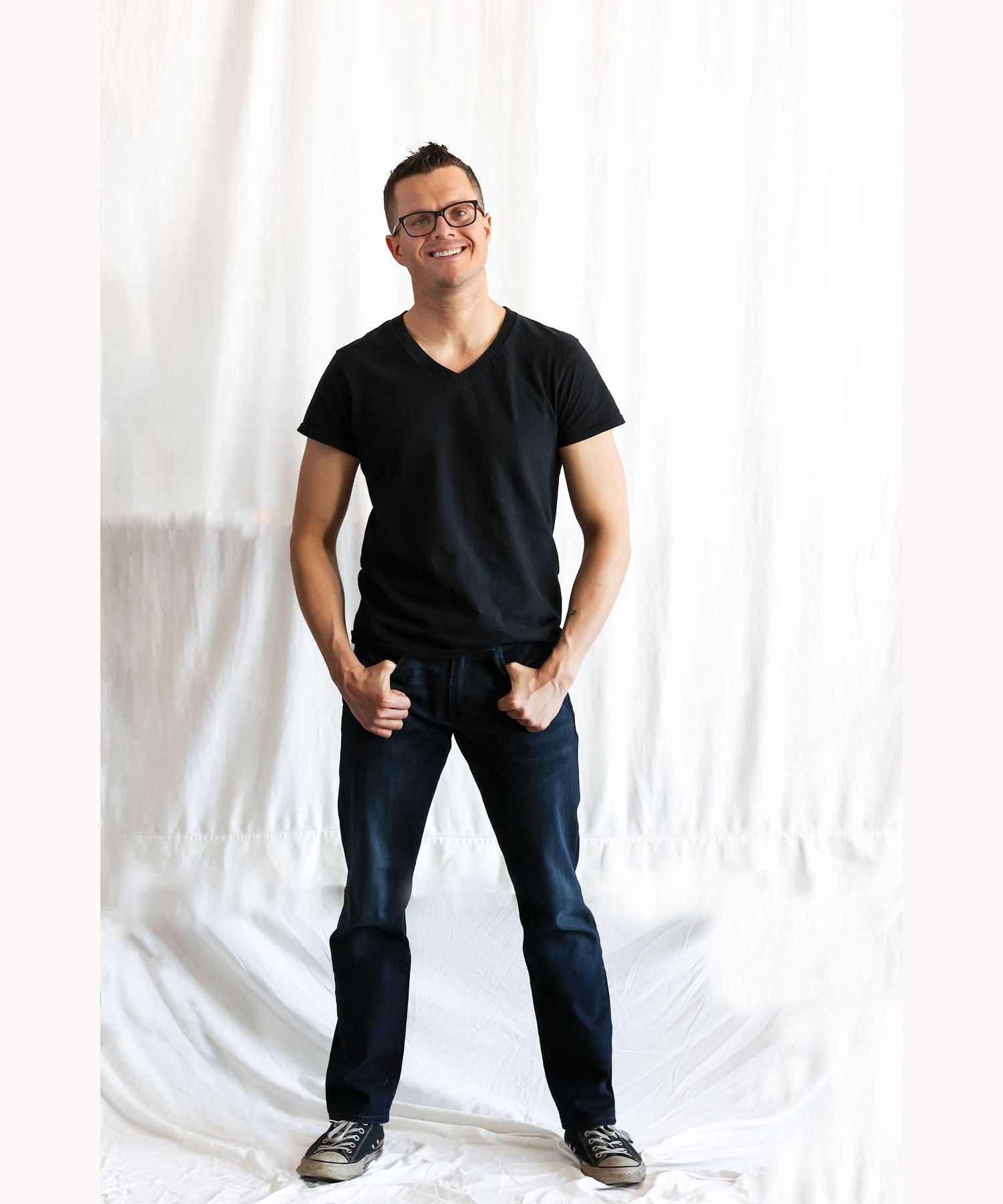 Smiling man in black v-neck shirt and dark jeans posing confidently against white fabric backdrop.