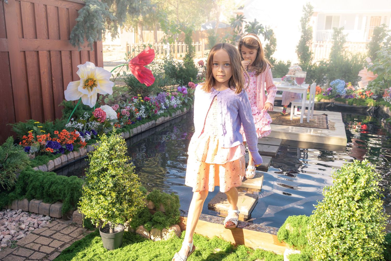 A girl in a pink skirt and lilac cardigan walks along stepping stones past giant paper flowers and blooms in a garden display.