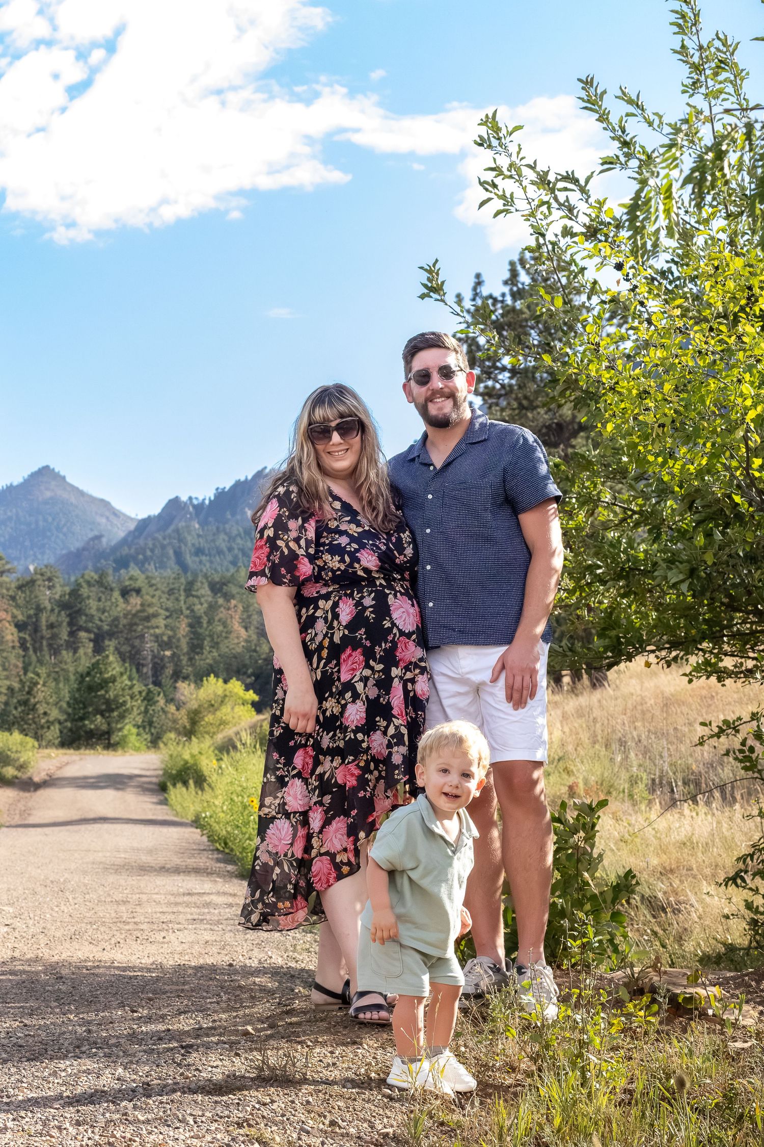 Family of three posing on a mountain trail, mother in floral dress, father in blue shirt, toddler in green, with peaks behind.