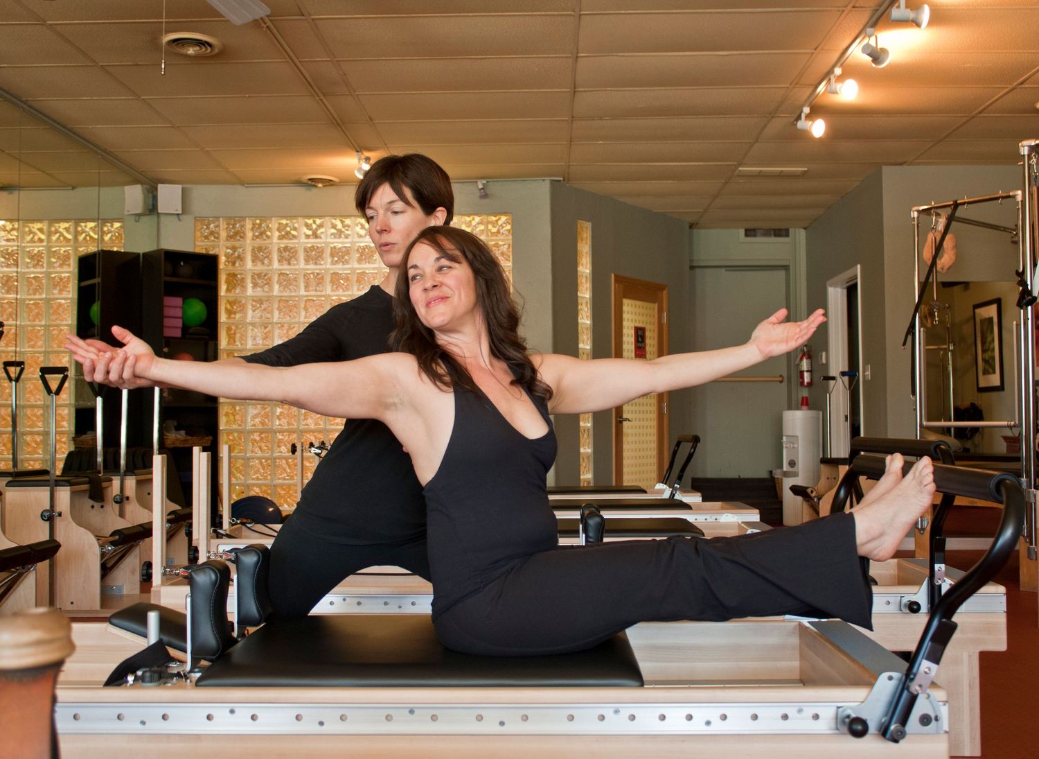 A Pilates instructor guides a client in a seated stretch on a reformer machine in a fitness studio.