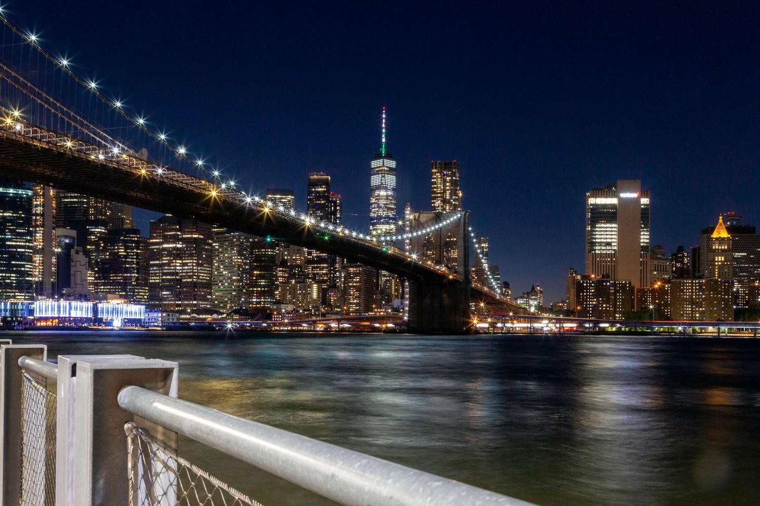 Brooklyn Bridge and Manhattan skyline illuminated at night reflecting on the East River.
