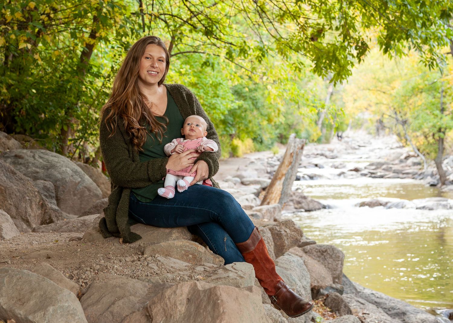 A smiling woman sits on rocks by a creek holding a baby, surrounded by lush green trees in a serene outdoor setting.